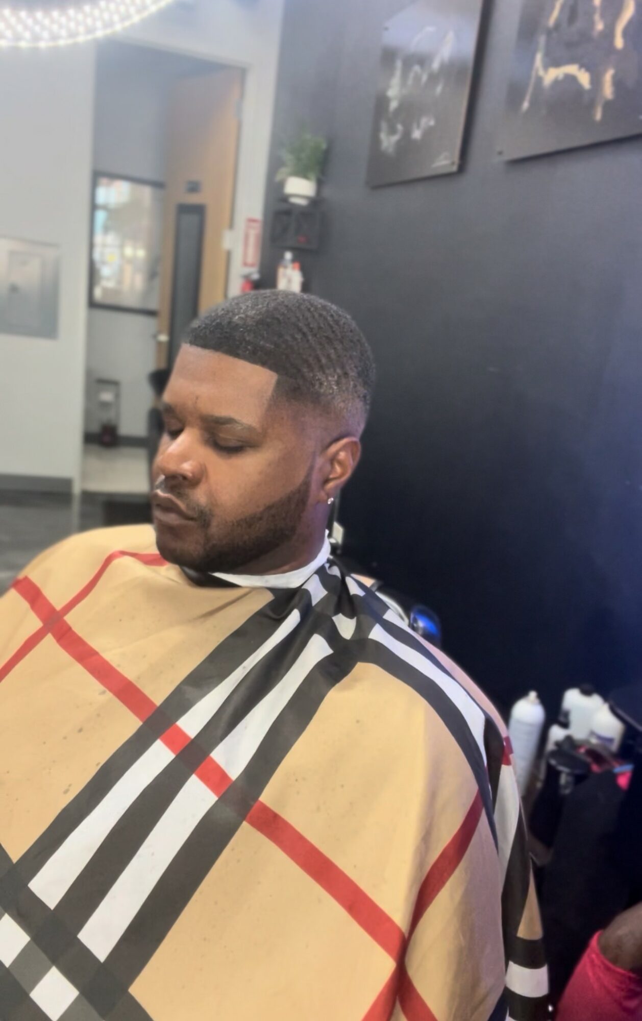 Man with short hair and beard sits with eyes closed, wearing a beige and black patterned cape, in a salon or barbershop.