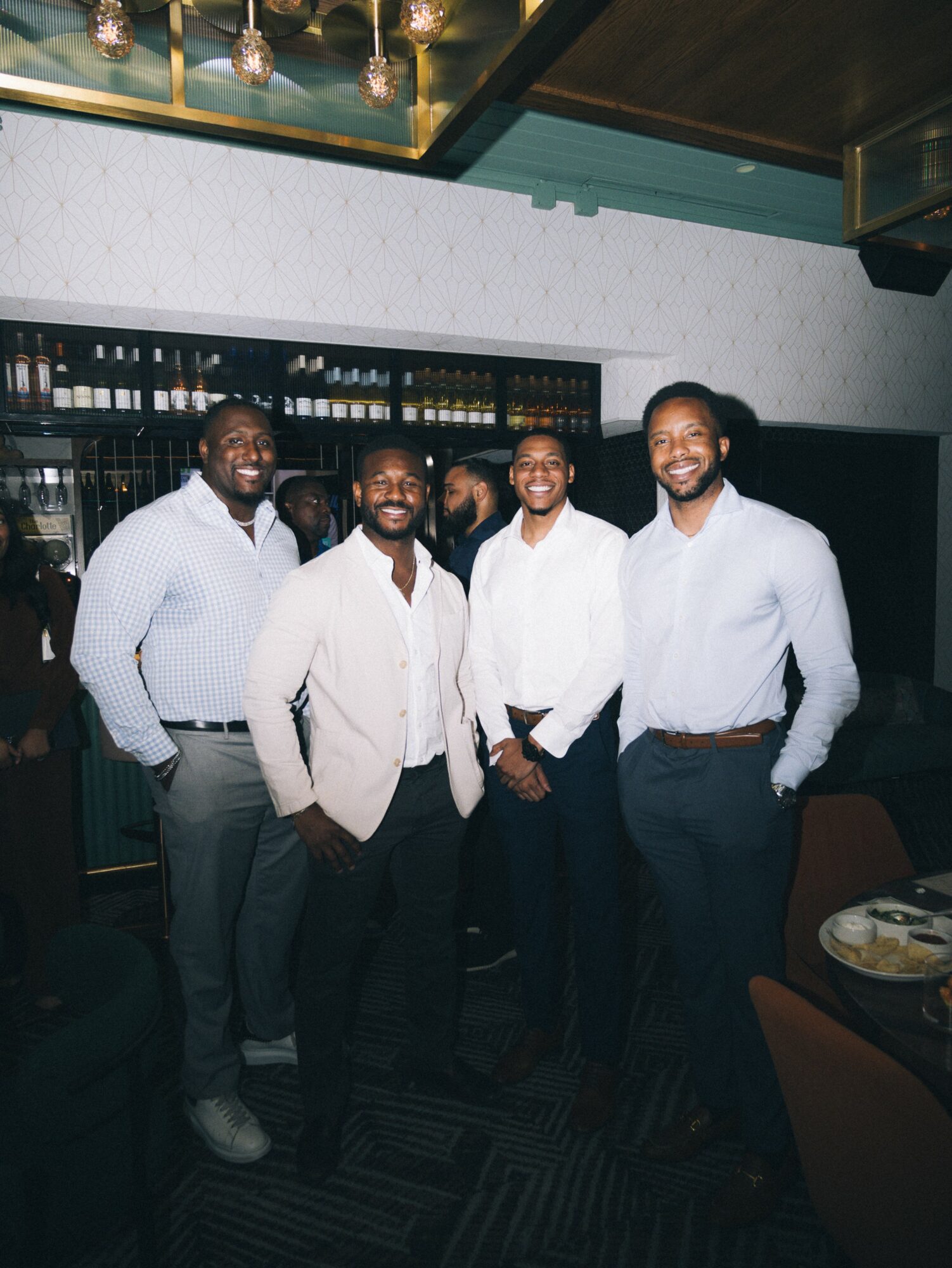 Four men stand together in a bar or restaurant, smiling, with shelves of bottles behind them.
