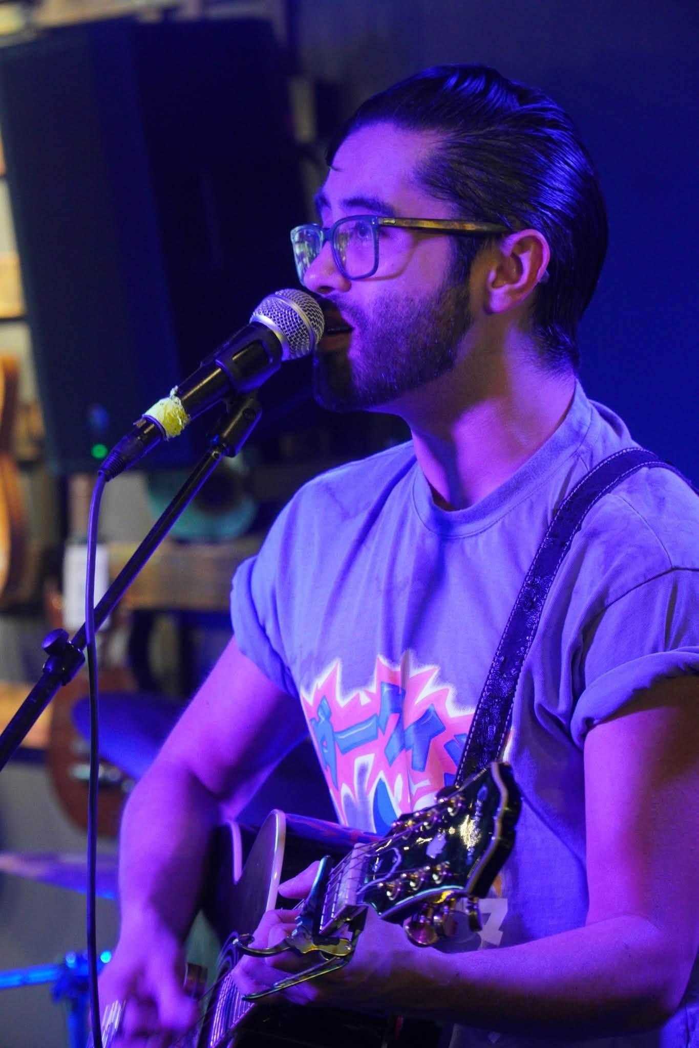 Person with glasses singing into a microphone while playing guitar, wearing a graphic t-shirt, in a dimly lit setting.