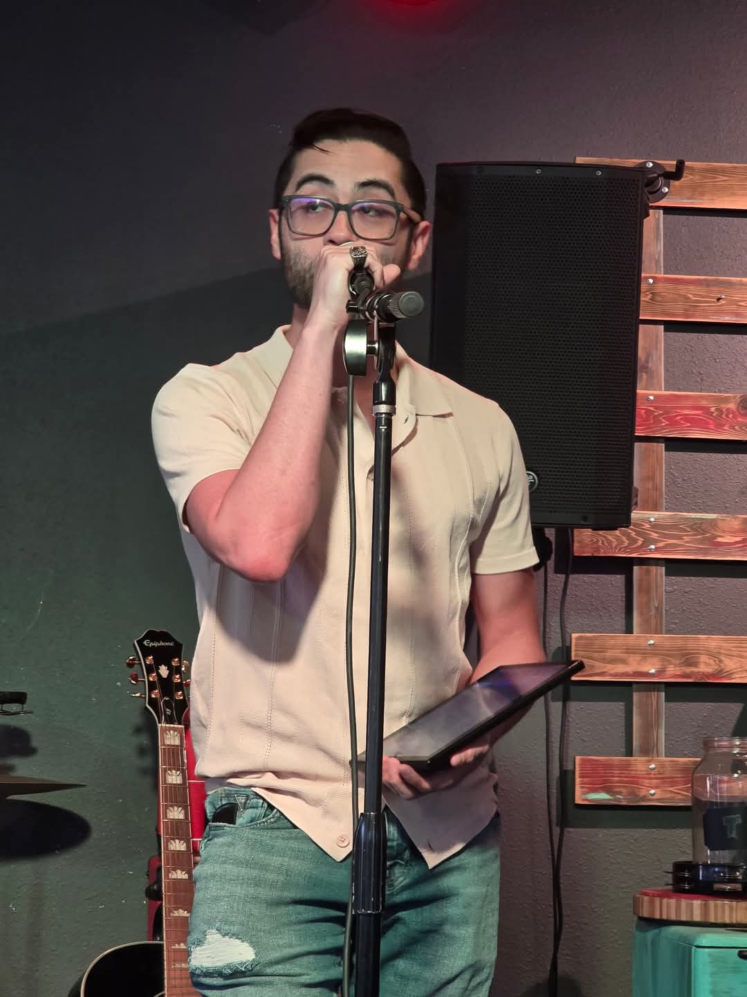 Person with glasses singing into a microphone, holding a tablet, on stage with a wooden backdrop.