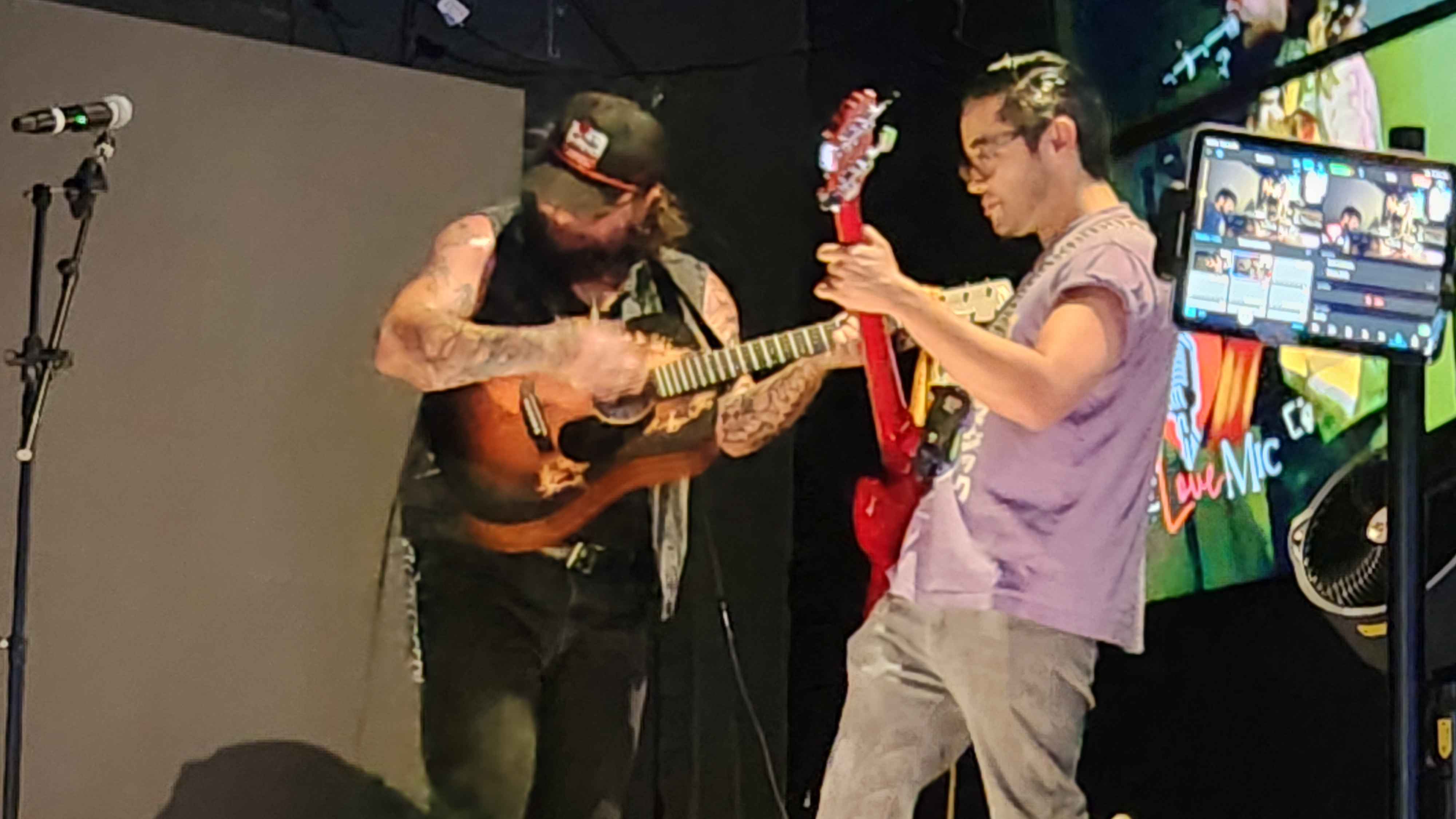 Two musicians playing guitars on stage, one with tattoos and a cap, the other with glasses and a purple shirt.