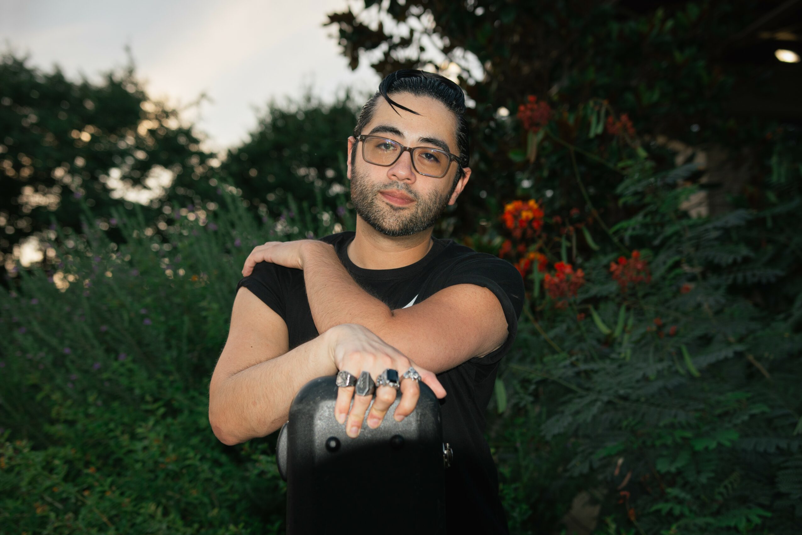 Young man with glasses and a beard, standing outdoors with greenery and flowers, looking at the camera.