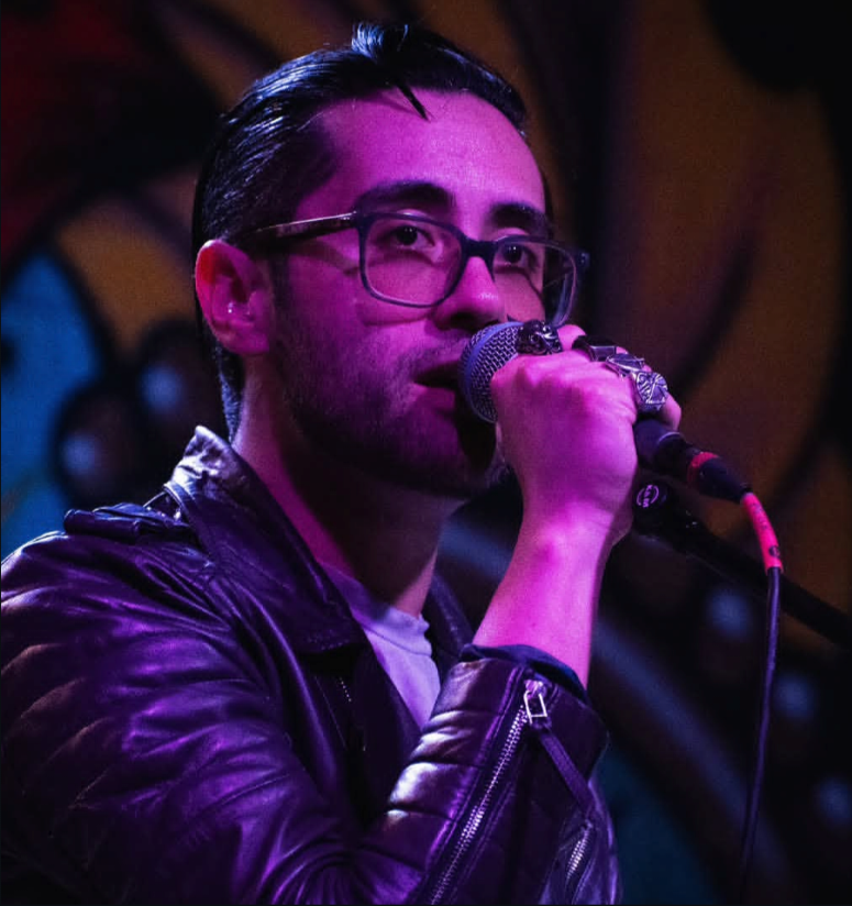 Person with glasses and dark hair holding a microphone, wearing a leather jacket, performing on stage.
