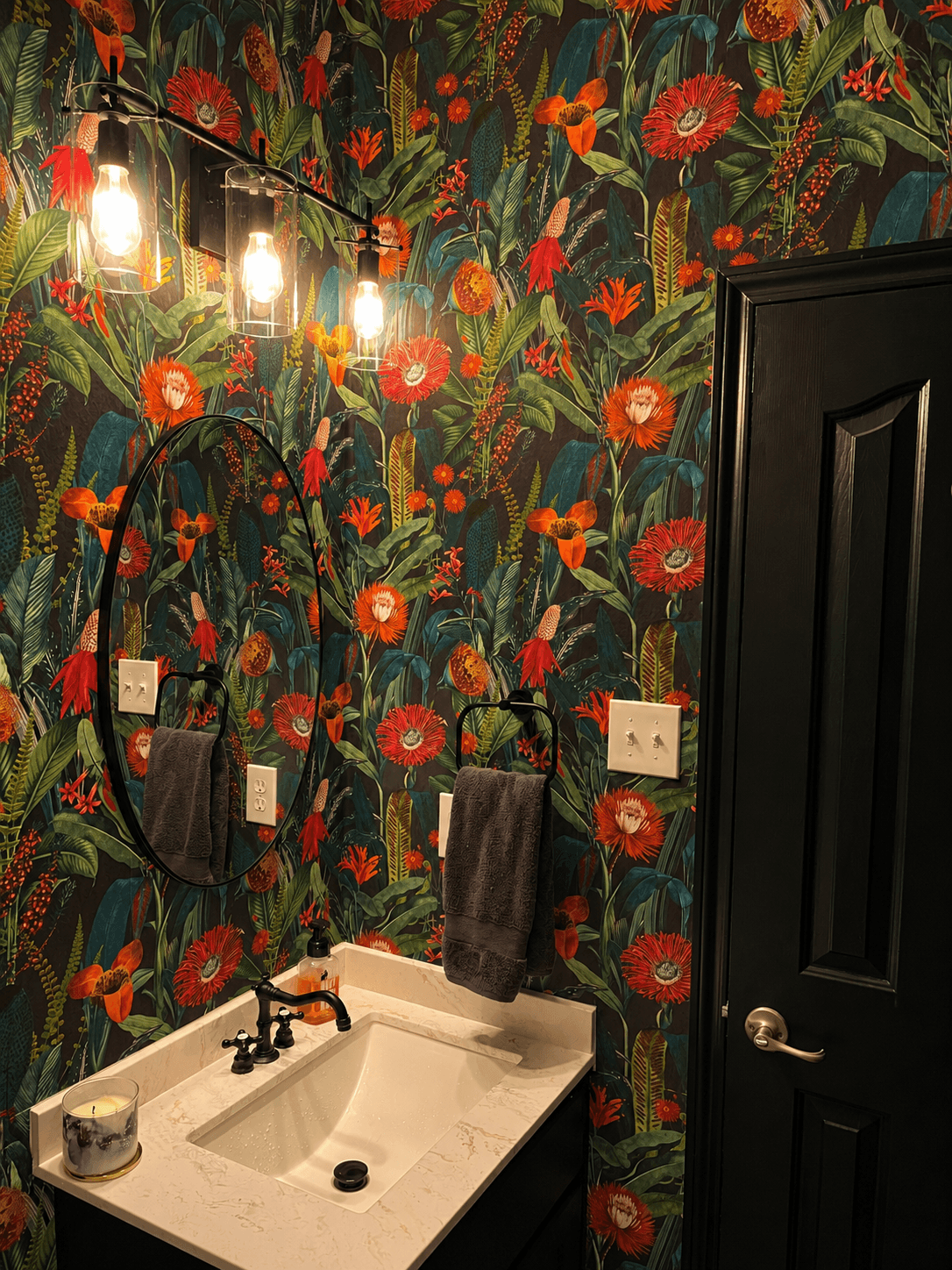 Bathroom with floral wallpaper, black mirror, white sink, towel, and black door, illuminated by hanging light bulbs.