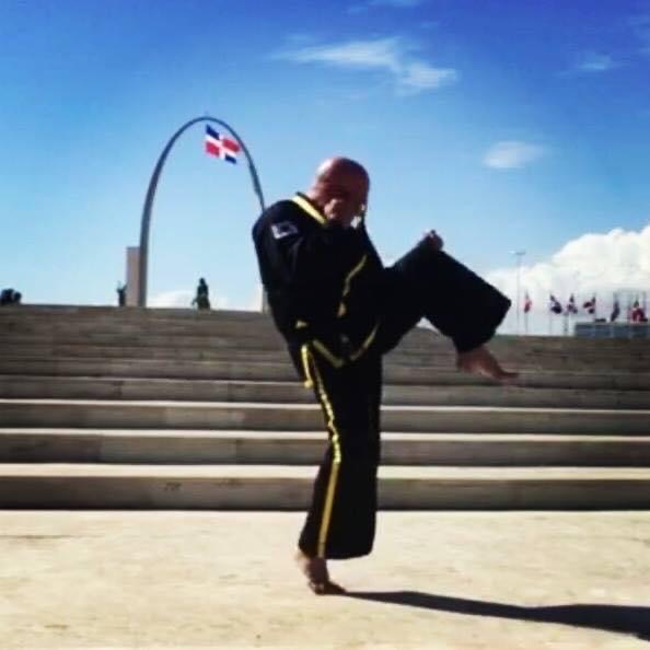 Person in martial arts uniform performing a high kick outdoors on steps, with a flag and blue sky in background.