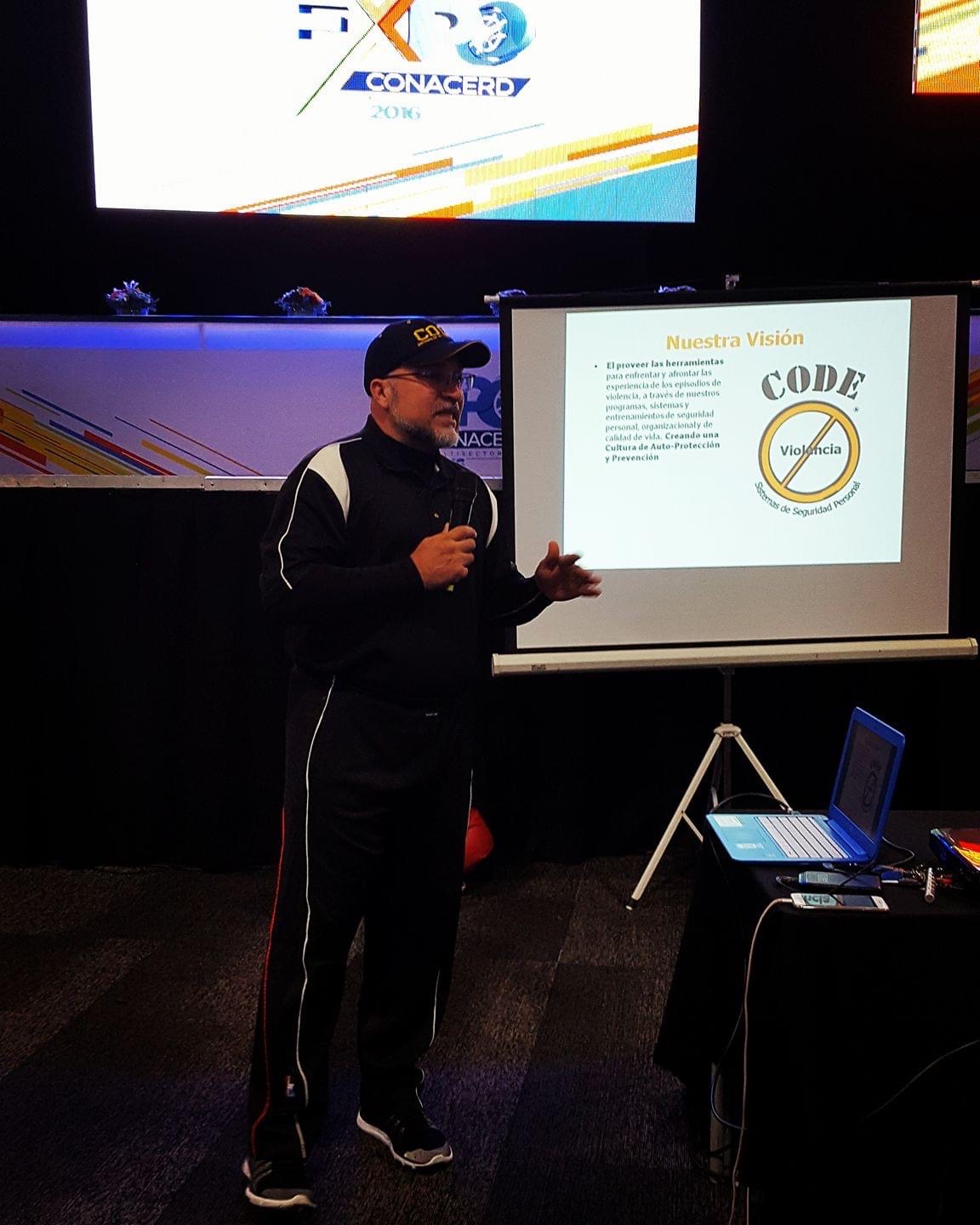 Person giving a presentation in front of a screen and a projector, with a laptop on a table nearby.