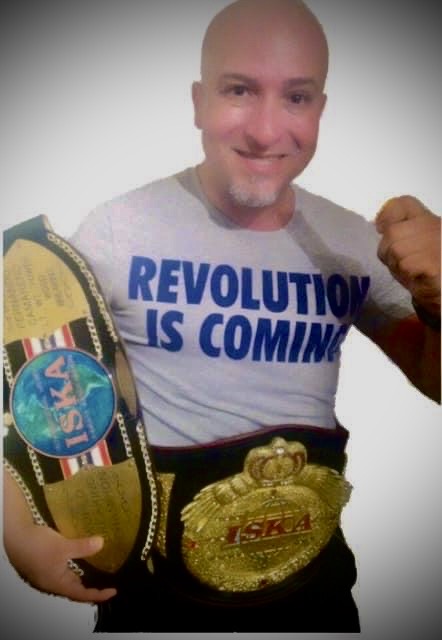Person with a bald head smiling, wearing a white T-shirt with blue text, holding championship belts, fist raised.