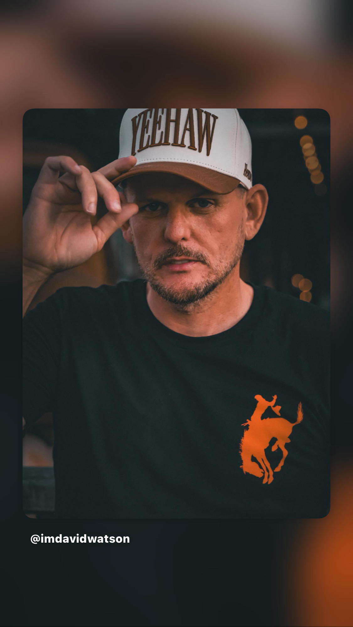 Man wearing a white cap with text, adjusting the cap with his right hand, wearing a black shirt with orange graphic, serious expression.