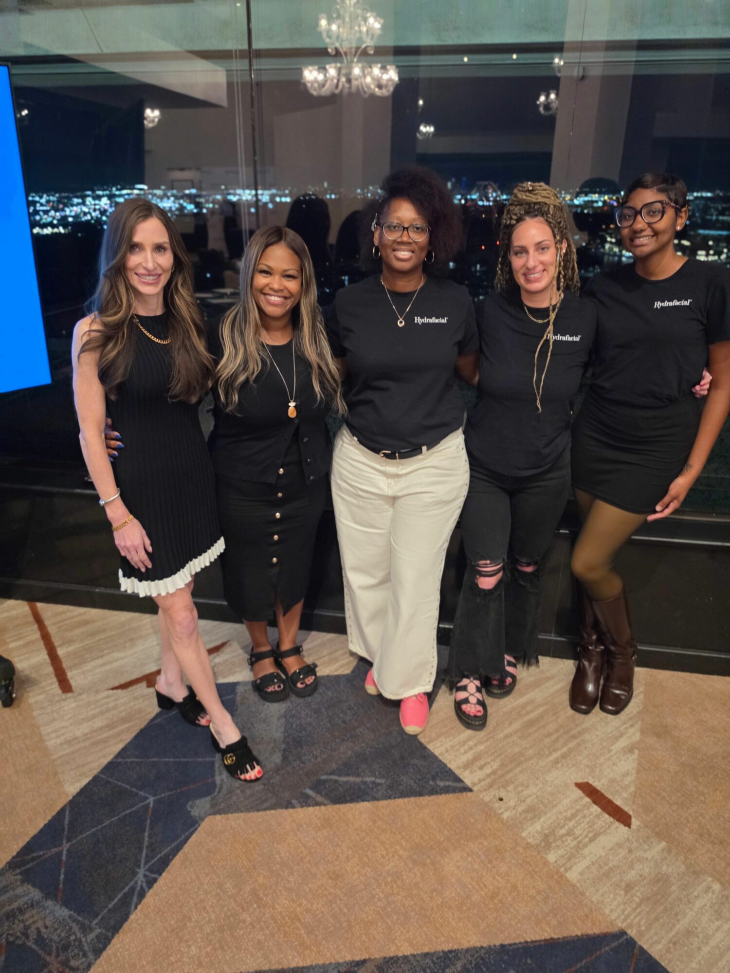 Group of five women standing together indoors, some smiling and posing, with a glass window and decorative flooring in the background.