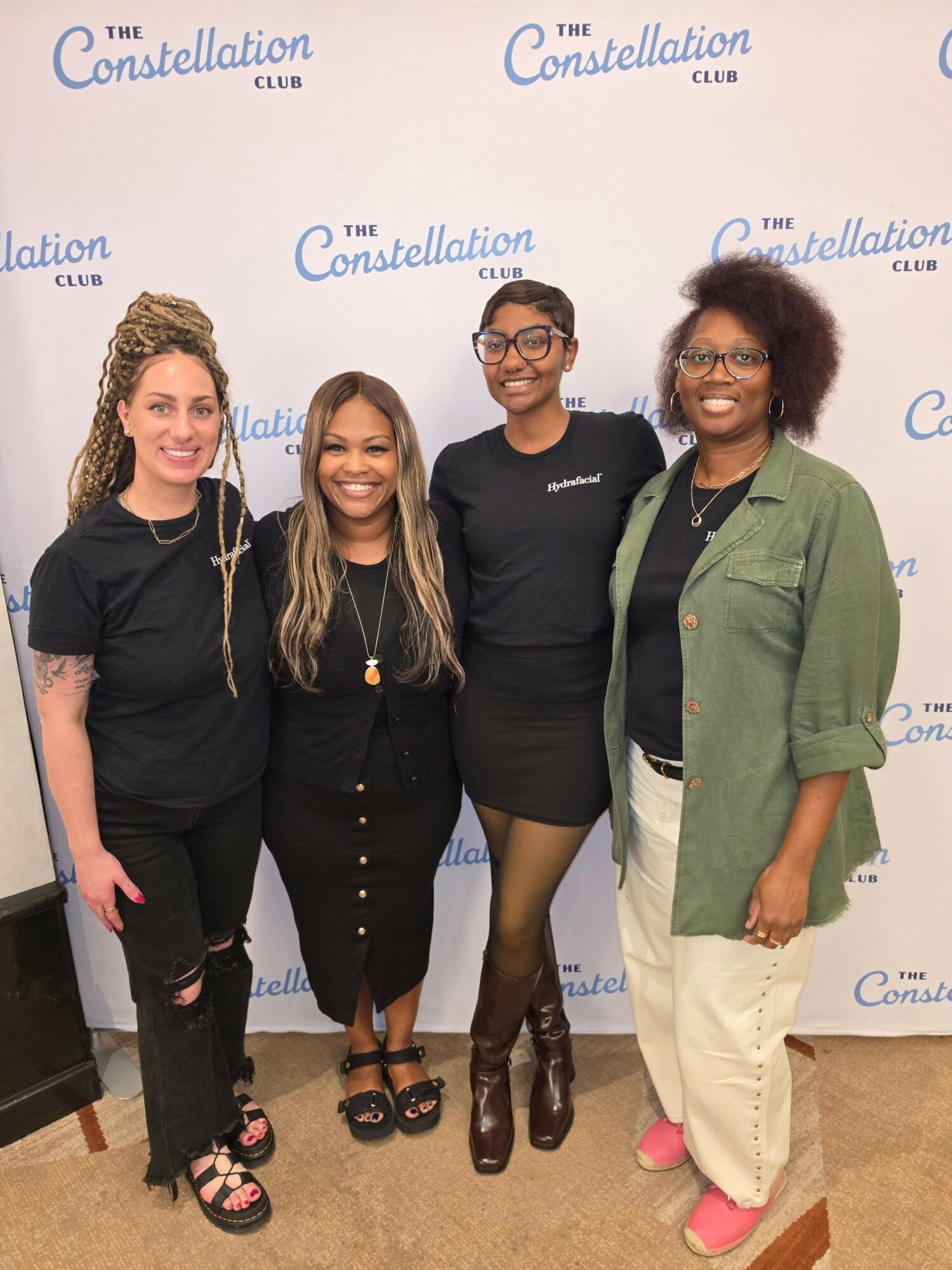 Four women standing together in front of a backdrop with text, smiling, dressed in black and green, with one wearing pink shoes.