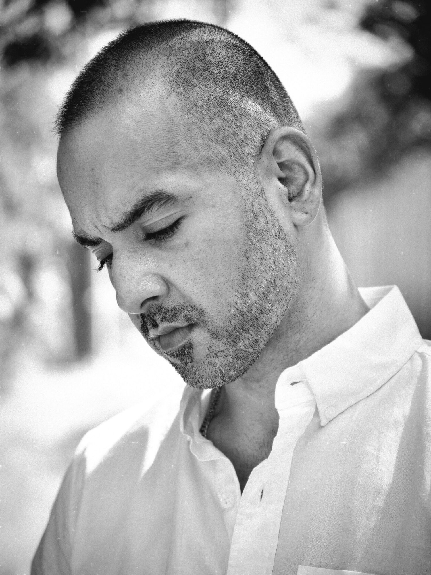 A man with a shaved head and beard looking down, wearing a white shirt, outdoors in black and white.
