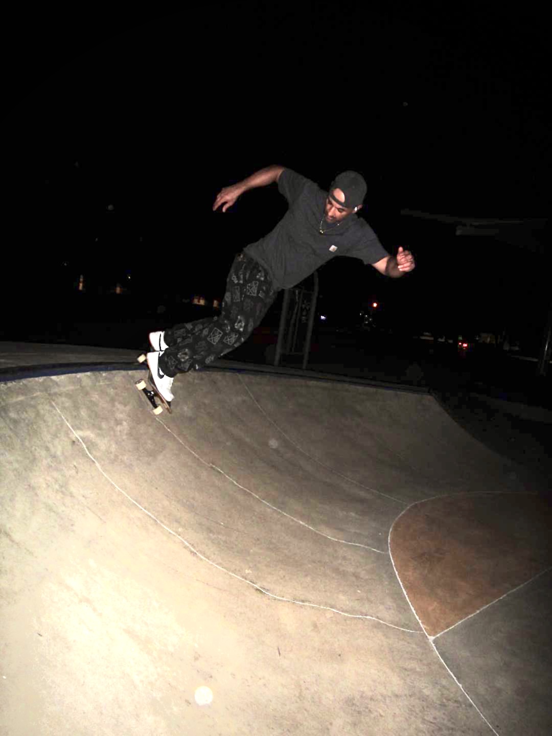 Skateboarder in dark clothing and cap performing a trick on a skatepark ramp at night.