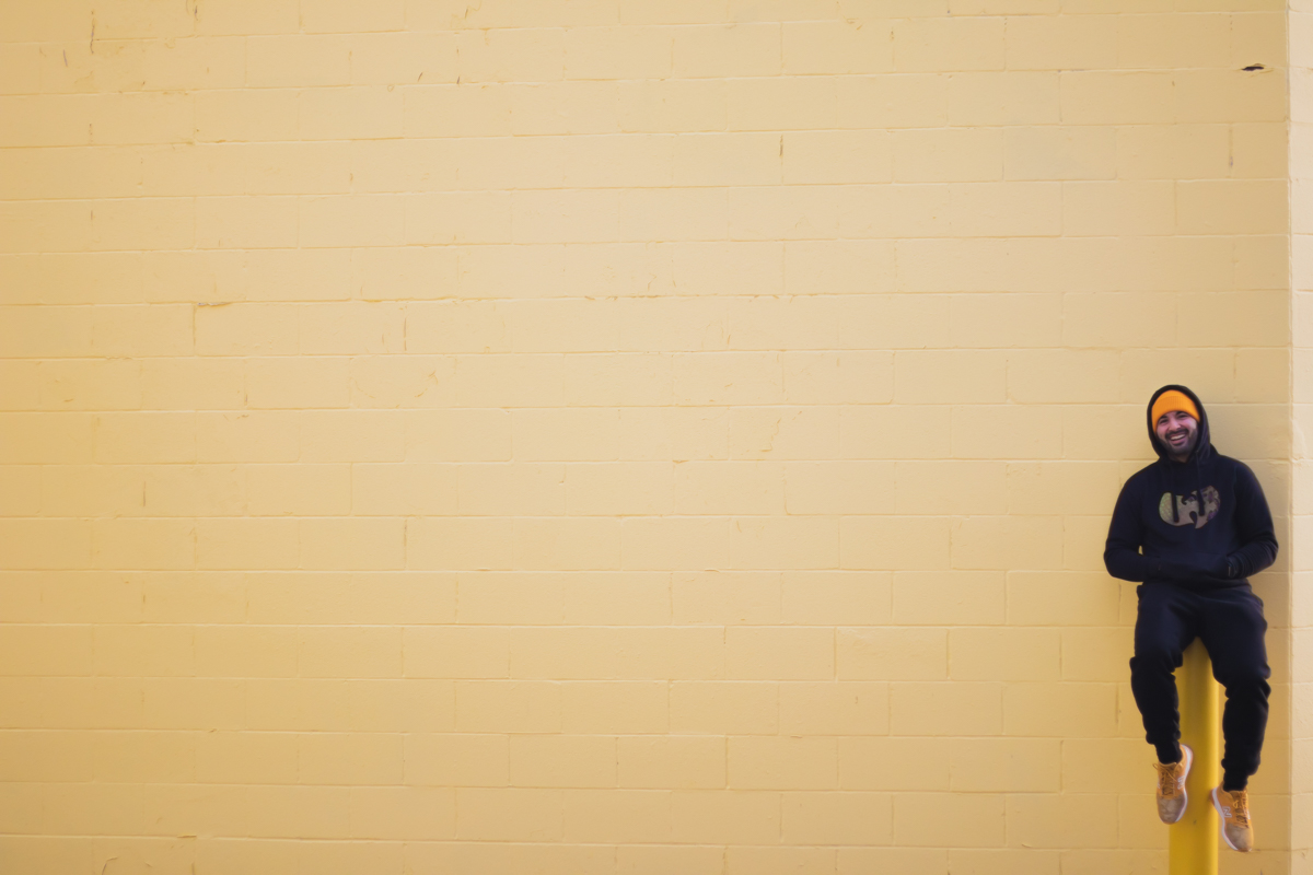 Person sitting on a yellow wall, smiling, wearing a dark hoodie and pants, with crossed arms and sneakers, on the right side.