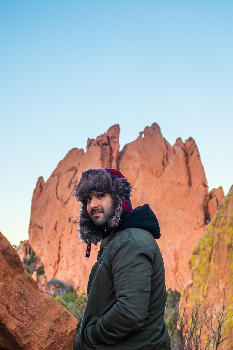 Man wearing a hat and jacket standing outdoors with rock formations and a clear blue sky in the background.