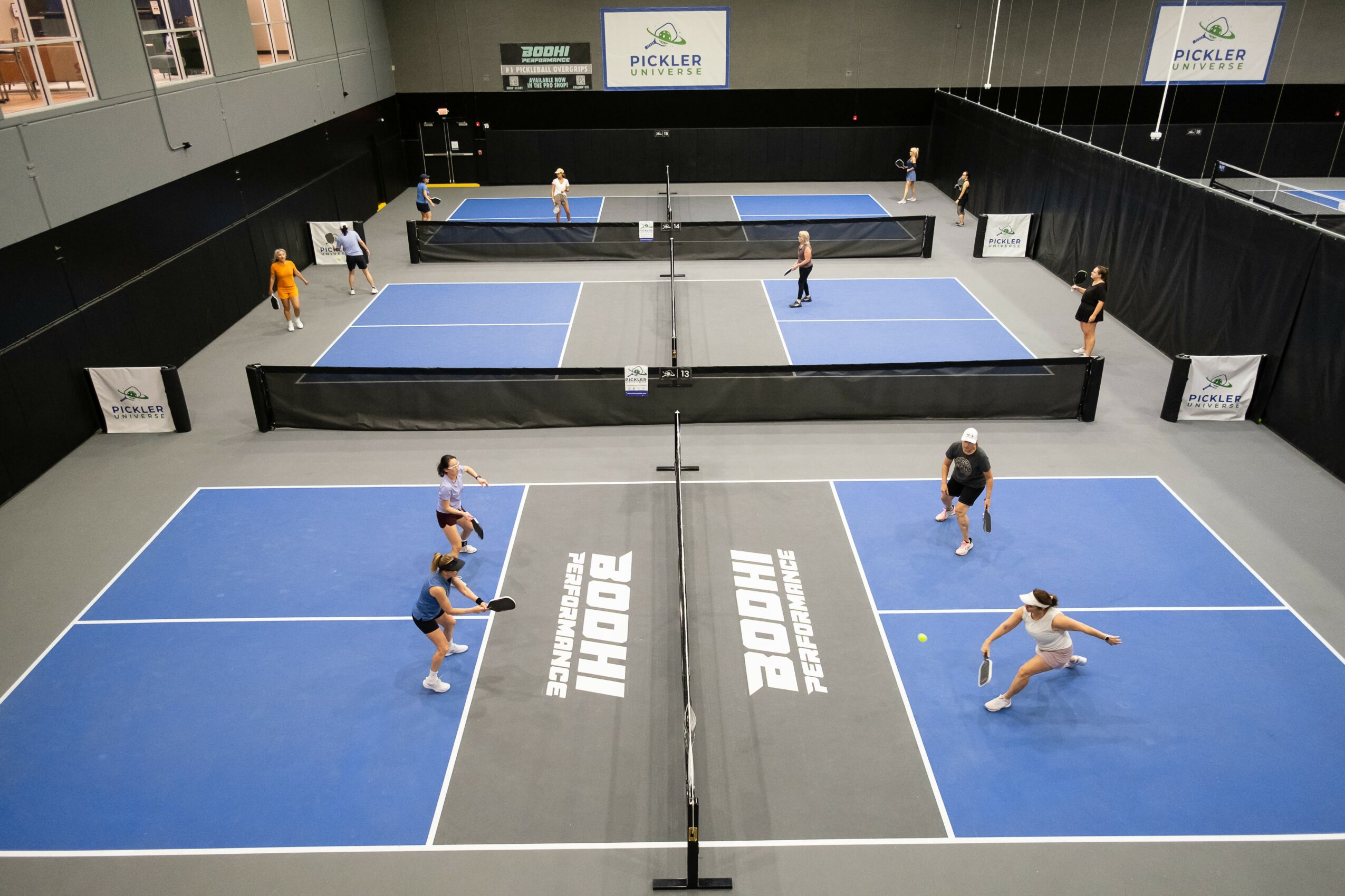 Indoor table tennis facility with four blue tables, players in action, black barriers, and signage, viewed from above.