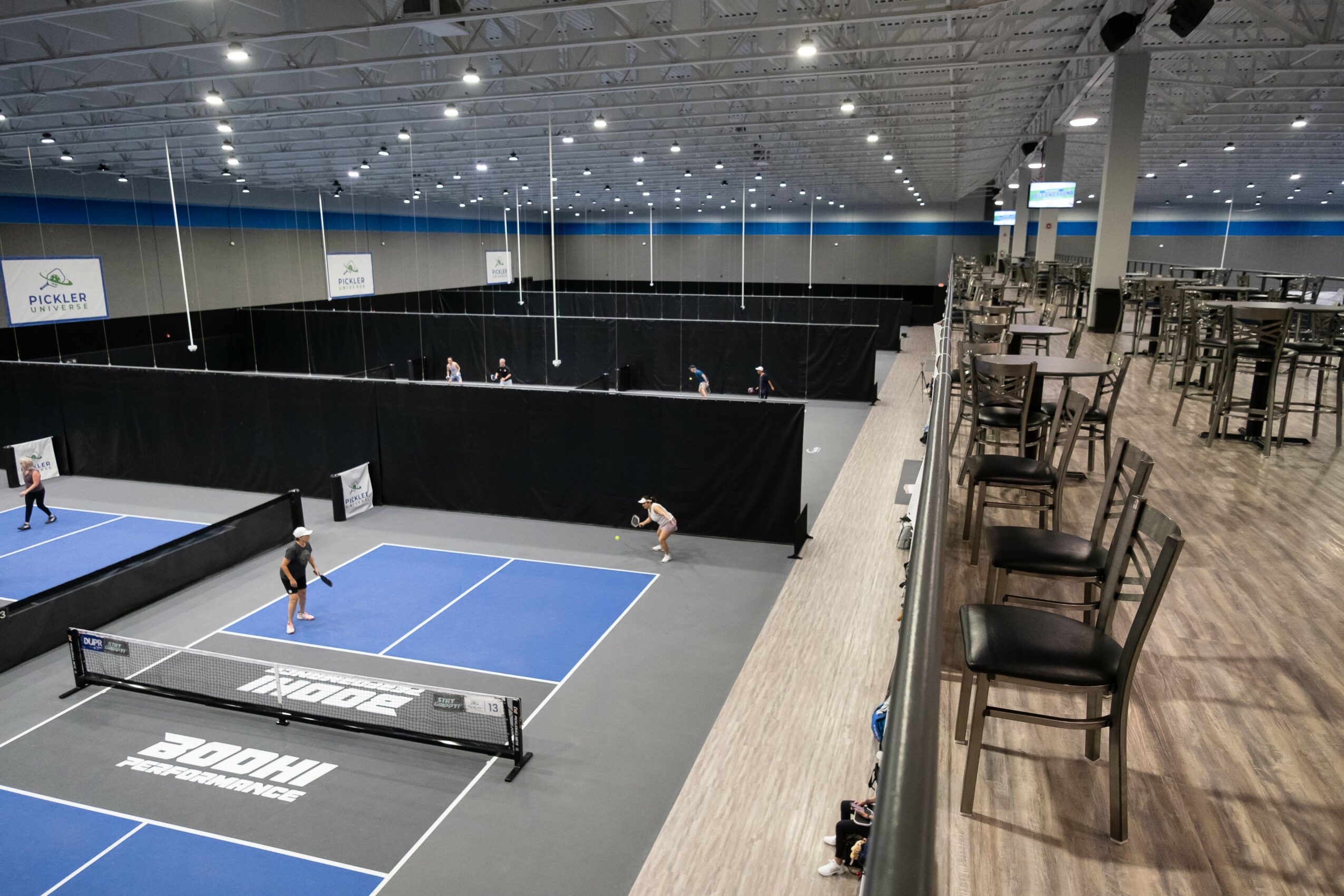 Indoor tennis court with players, seating area with chairs, and a high ceiling with lights.