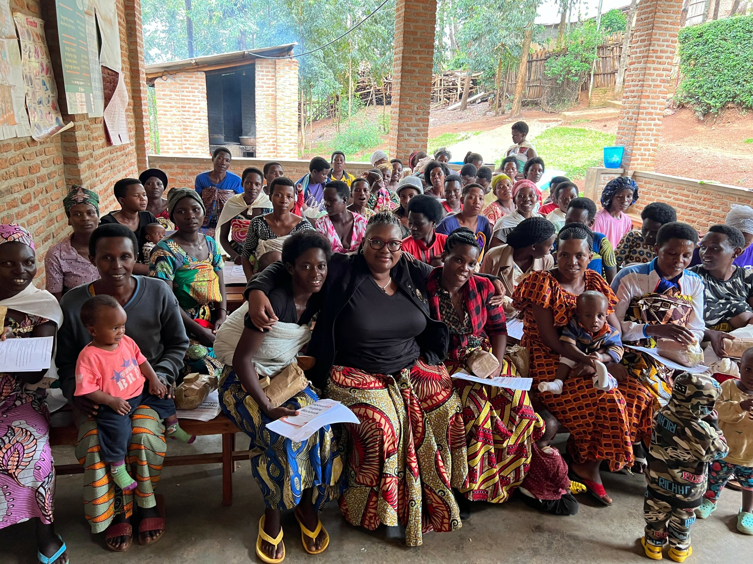 Group of people, including children and adults, sitting outdoors under a covered area with trees and brick structures in background.
