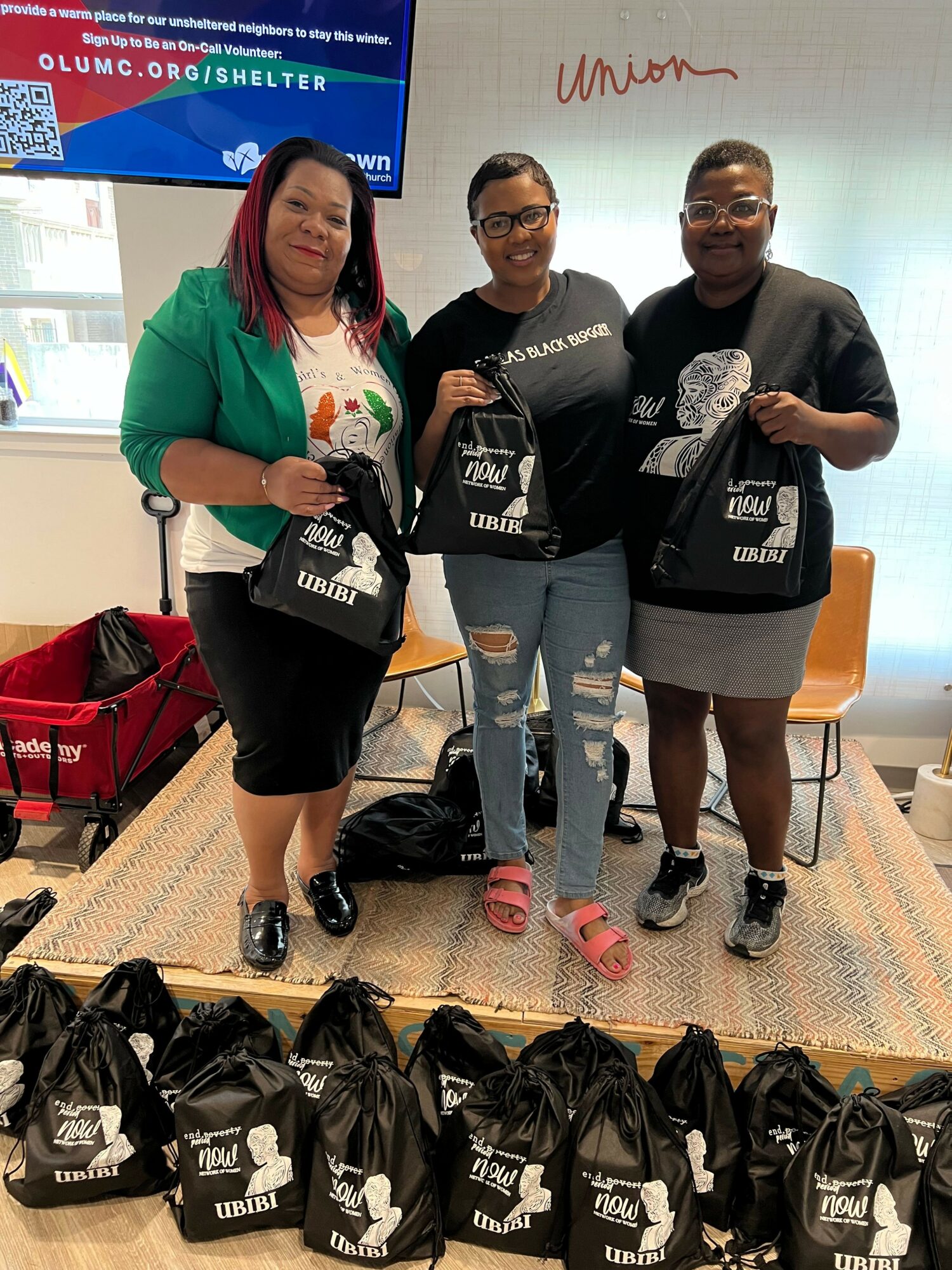 Four people standing indoors, holding black bags with white text, with a woman in a green jacket in front, and bags on the floor.