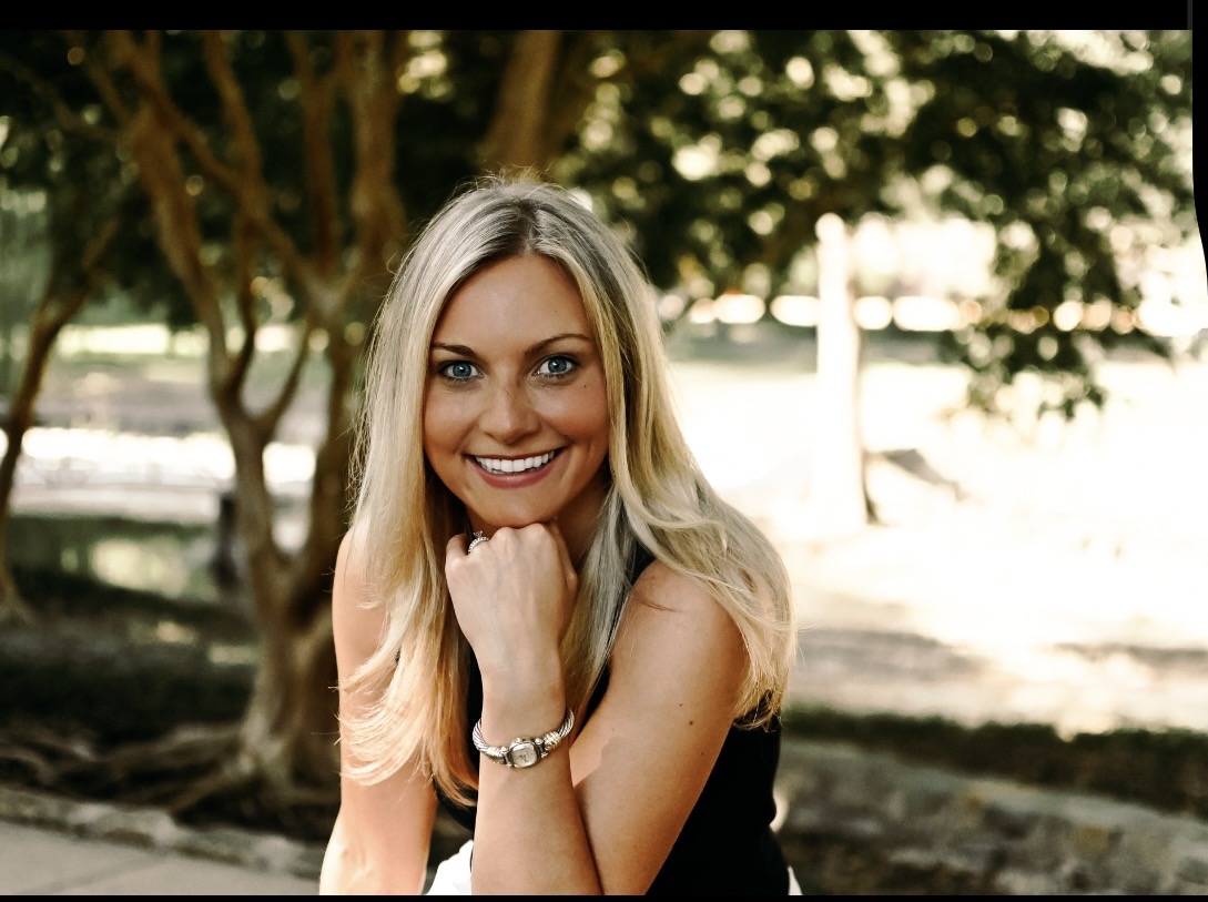 Smiling woman with long blonde hair outdoors, resting her chin on her hand, trees in background.