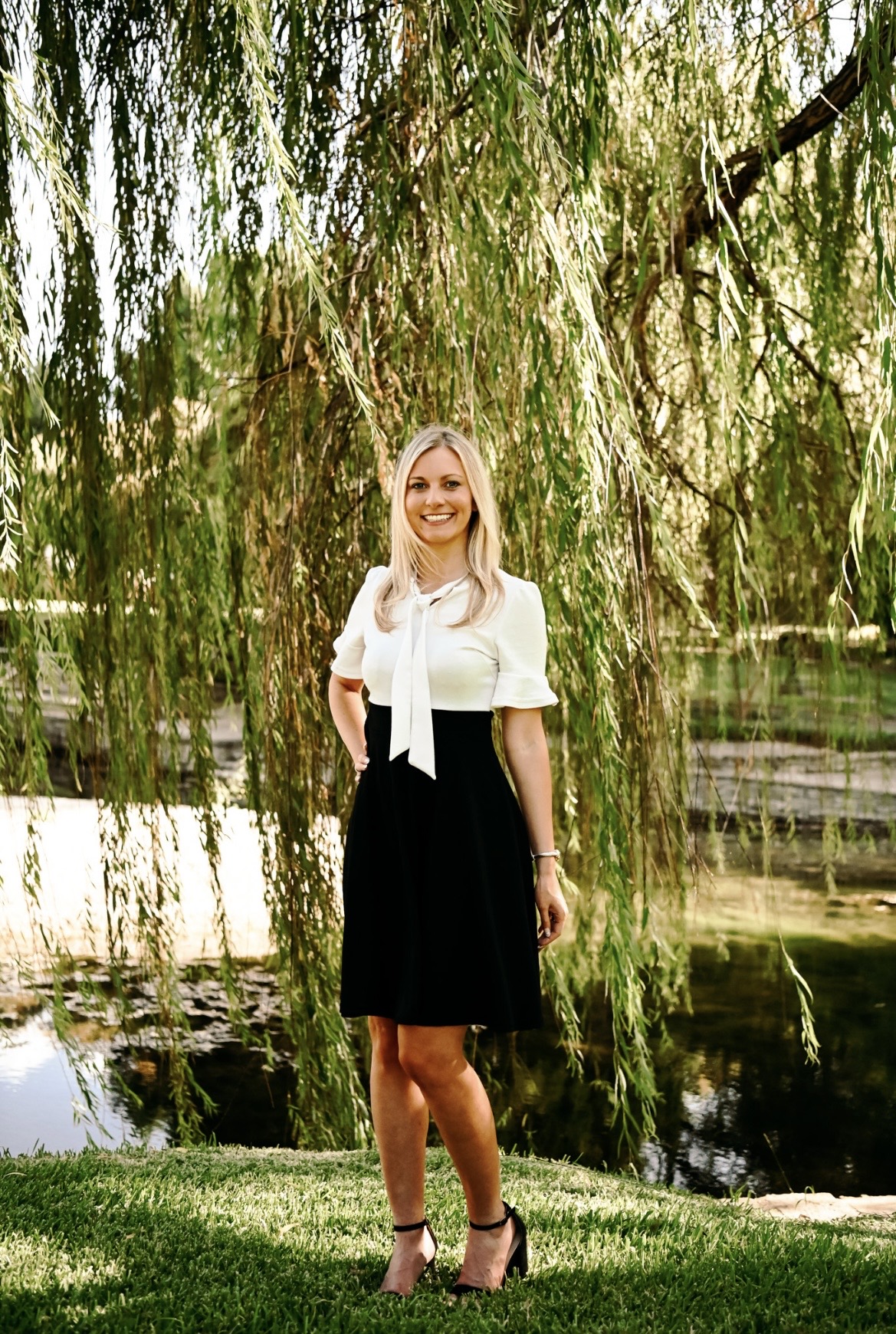 Woman standing outdoors near water with hanging tree branches, smiling, wearing a white top and black skirt.
