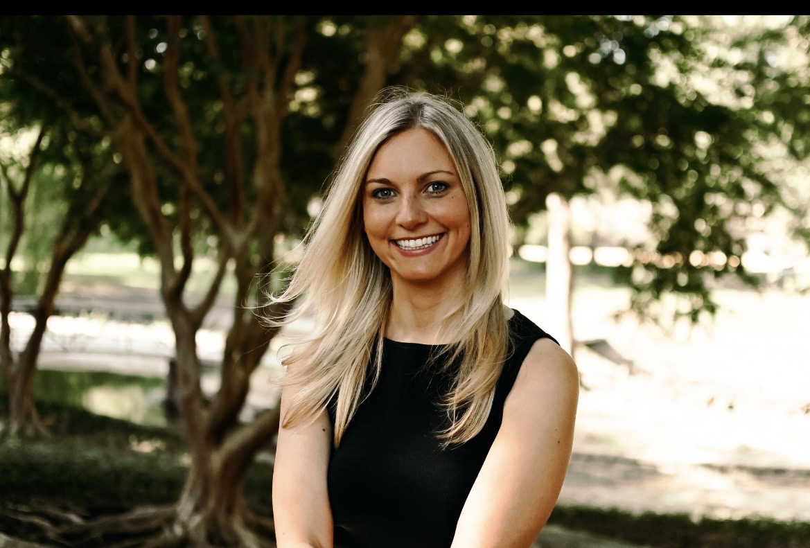 Smiling woman with long blonde hair outdoors, trees and sunlight in background.