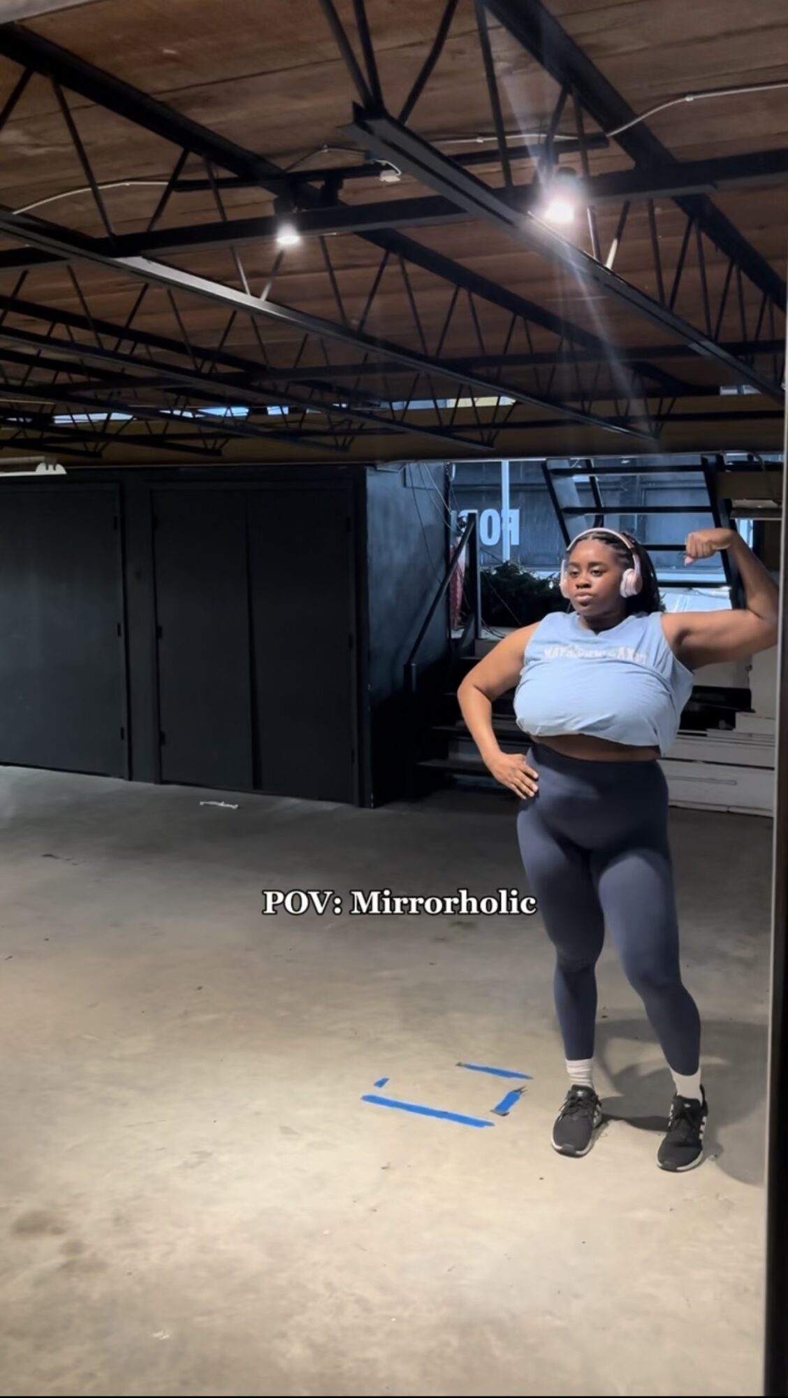 Woman taking mirror selfie in gym, flexing arm, wearing light blue top and dark leggings, with gym equipment in background.