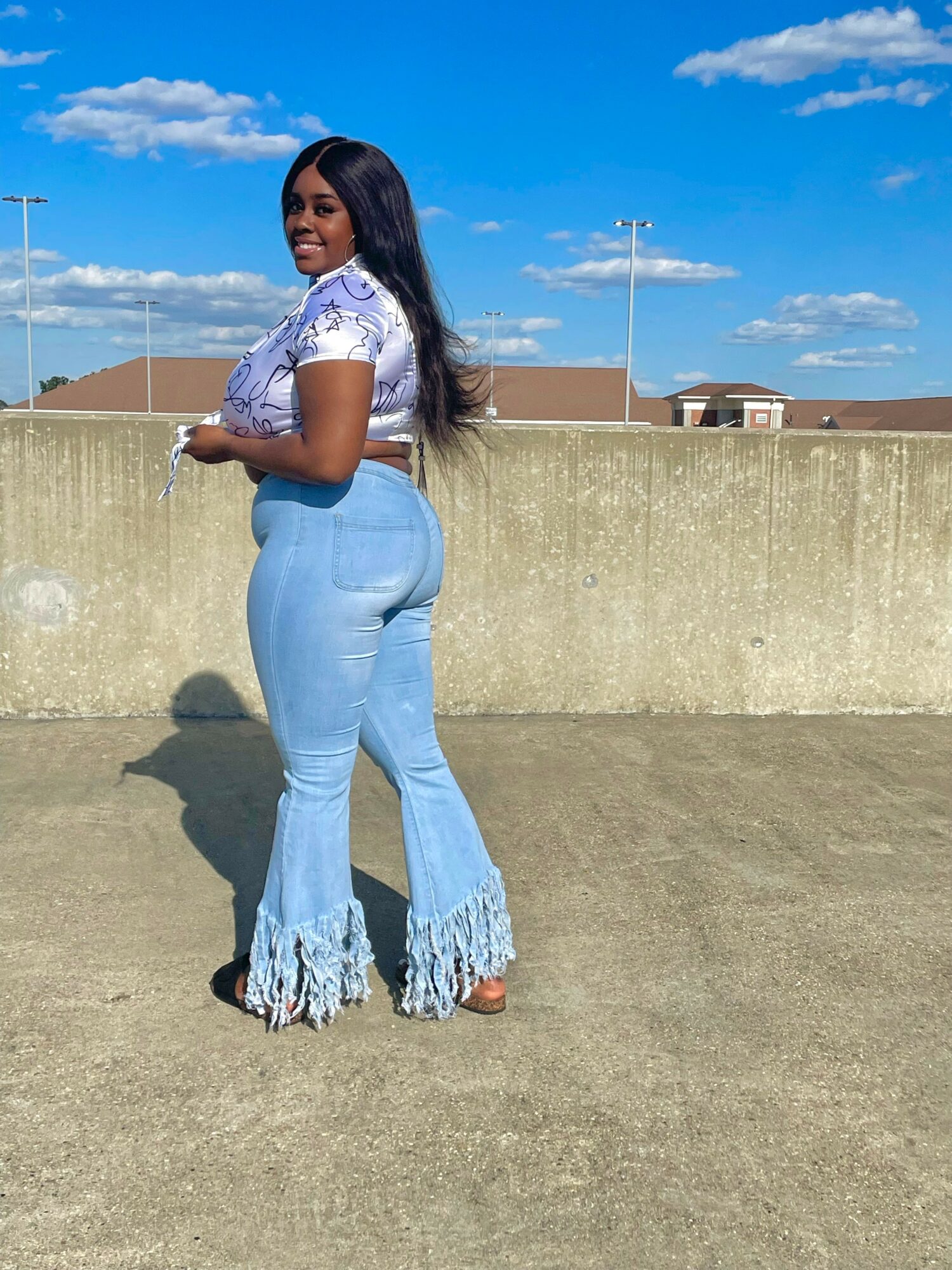 Woman standing outdoors against a concrete wall, wearing a white top and light blue jeans with ruffled hems, smiling.