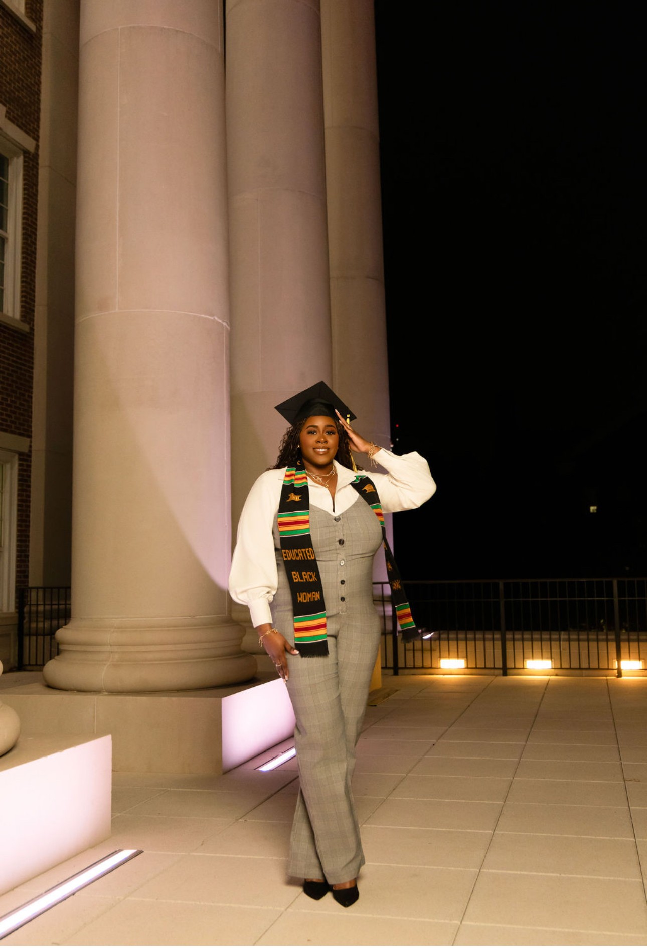 Graduate in cap and gown standing outdoors at night near large columns, smiling and saluting.