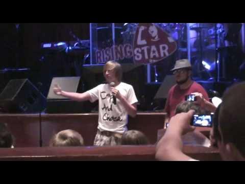 Child speaking on stage with two adults, audience watching, stage with sign and lights in background.