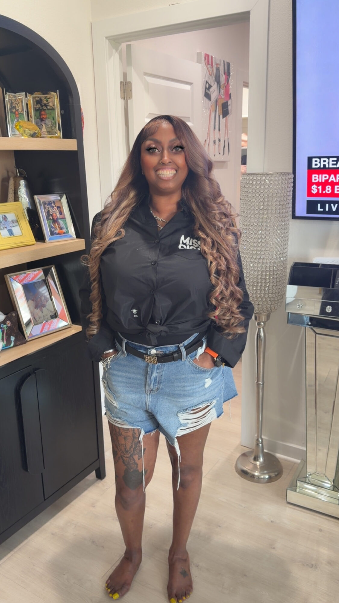 Smiling woman with long hair in a black jacket and denim shorts standing indoors near a bookshelf.