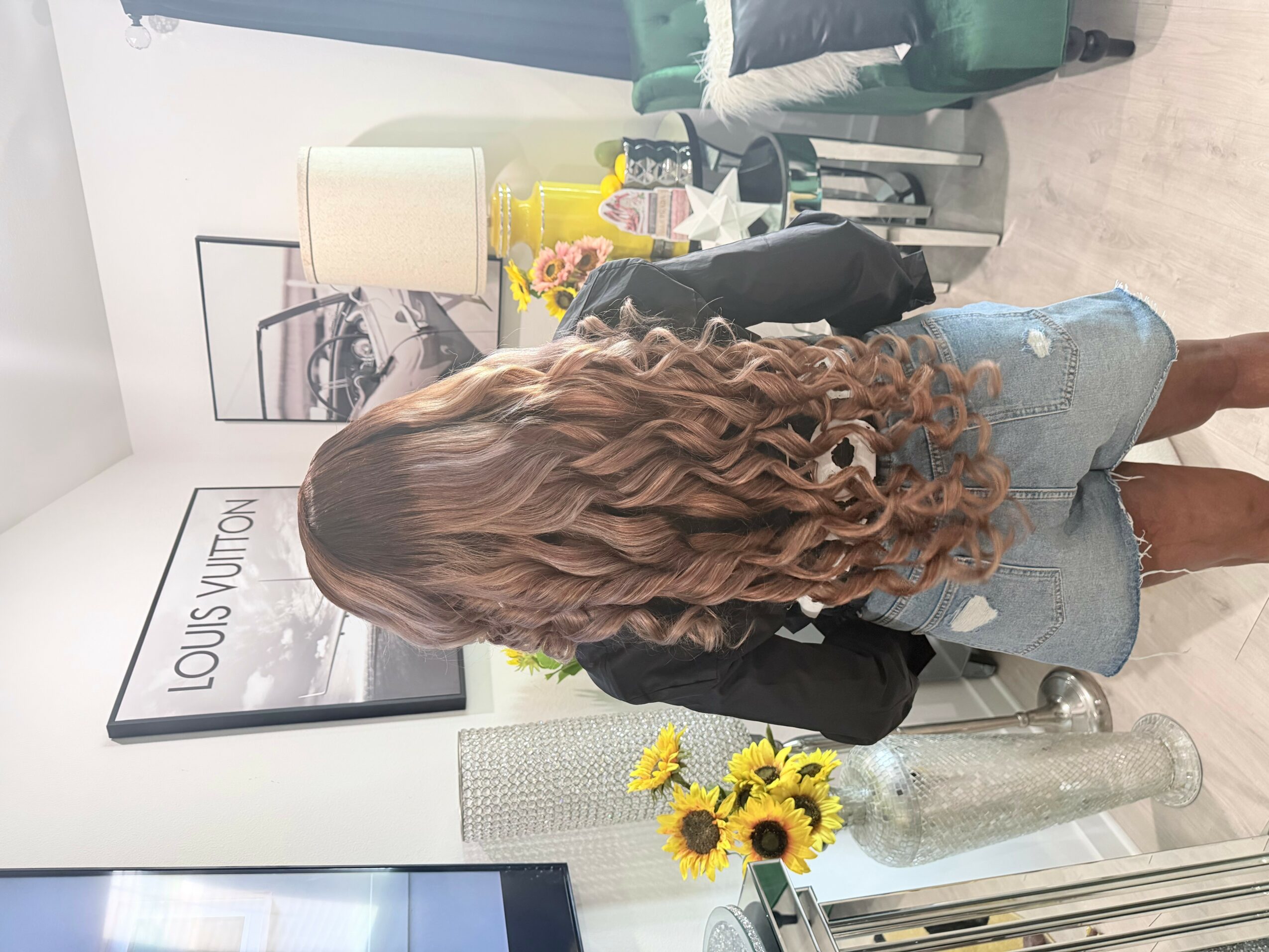 Person with long, curly hair standing in a room with a framed Louis Vuitton poster, a glass table, and a bouquet of sunflowers.