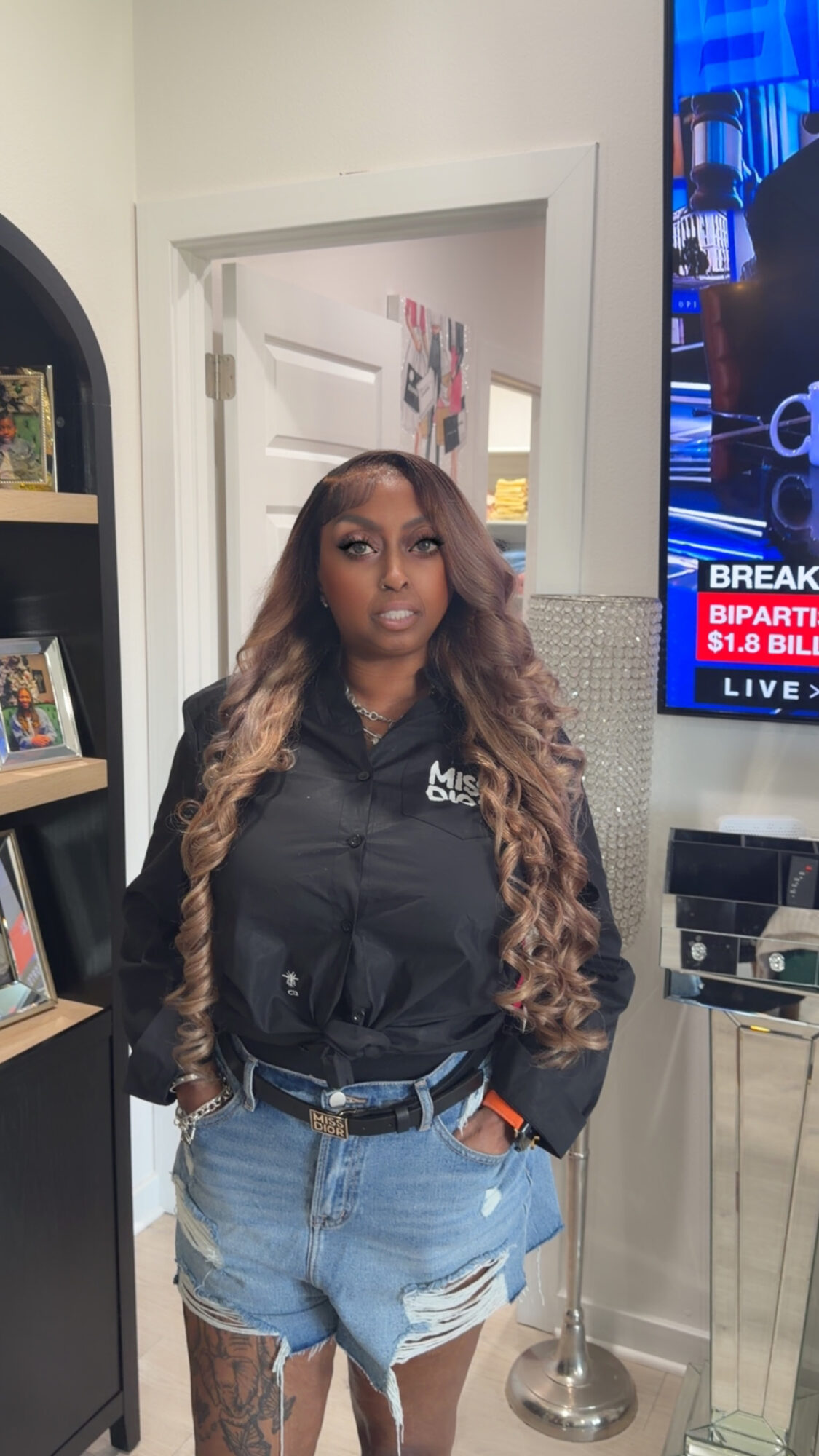 Woman with long wavy hair wearing a black jacket and denim shorts, standing indoors near a TV and bookshelf.