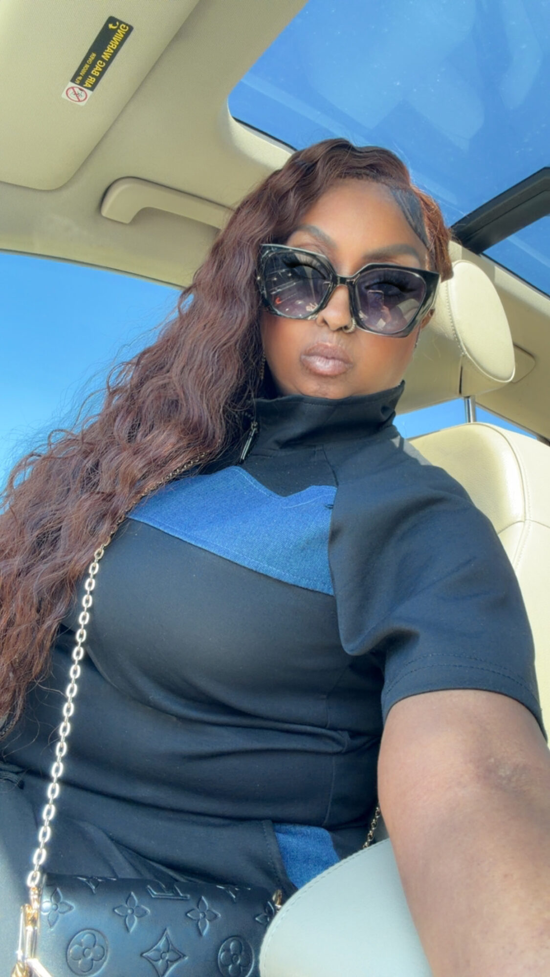 Woman with long wavy hair wearing sunglasses and a black and blue top, sitting in a car with a sunroof open.