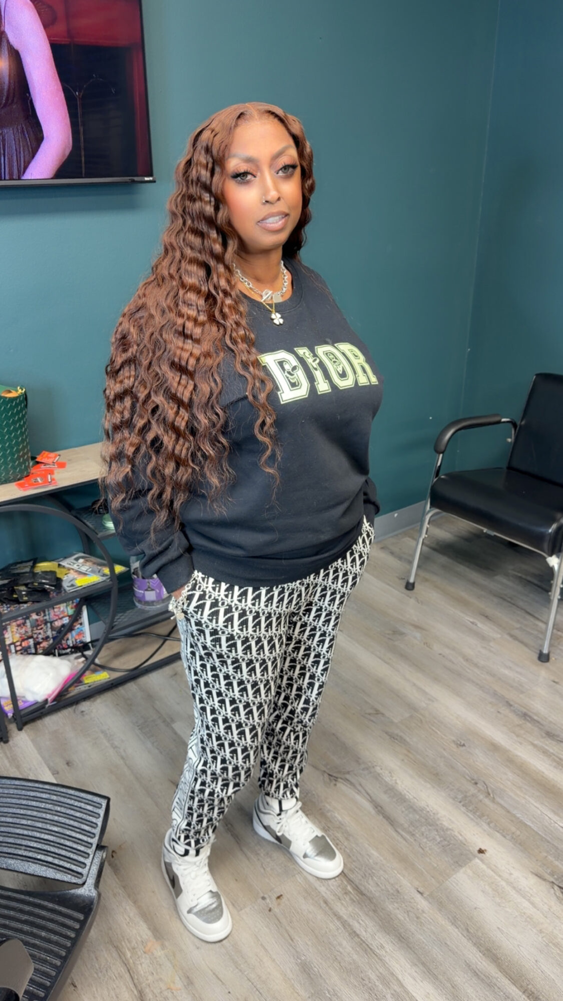 Woman with long curly hair standing indoors, wearing a black sweatshirt and patterned pants, looking at the camera.