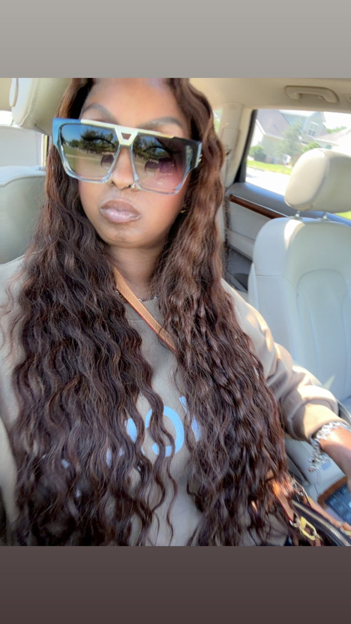 Woman with long wavy hair wearing large sunglasses in car, sunlight outside, neutral expression.
