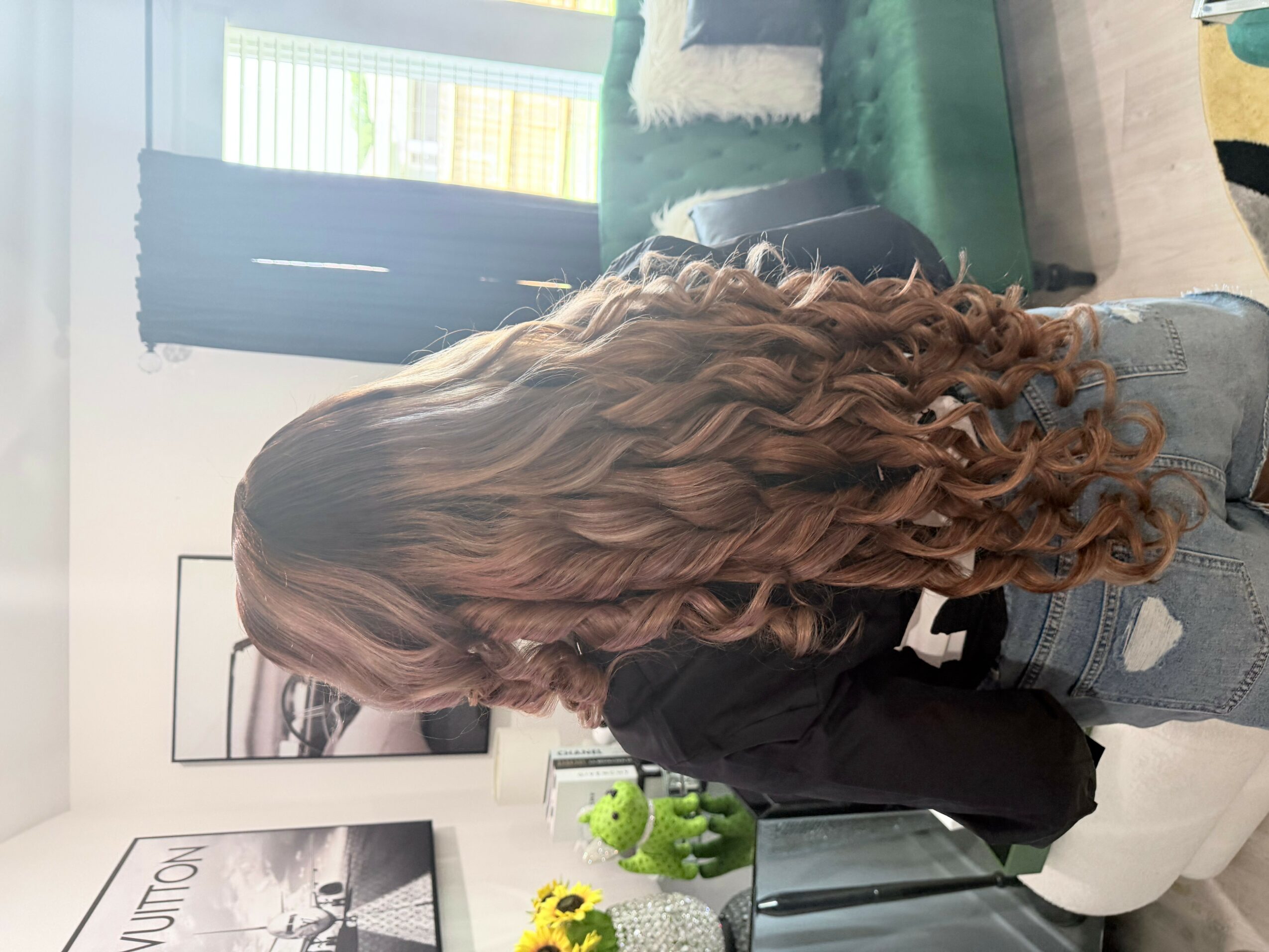 Person with long, curly hair sitting on a white chair in a room with a green sofa and window, facing left.