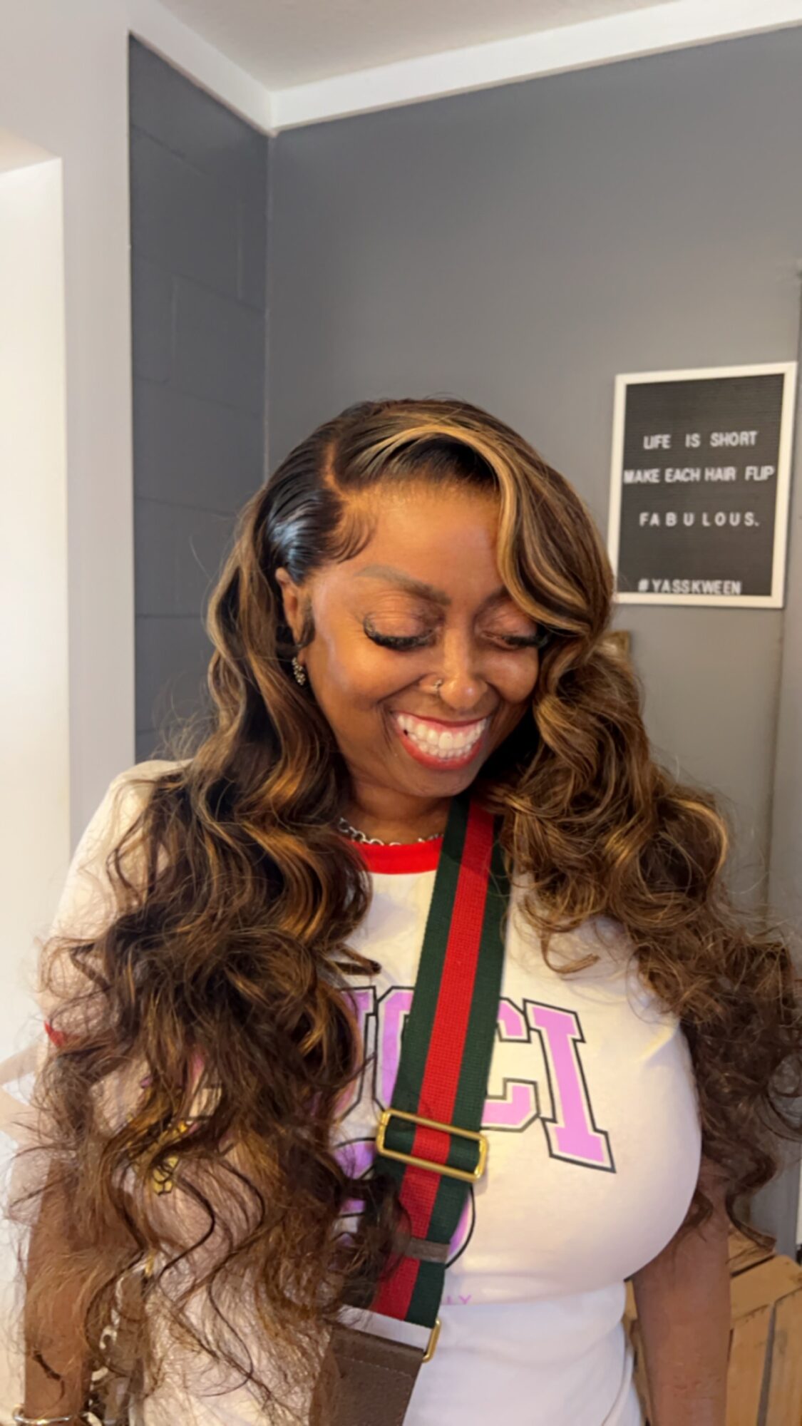 Woman with long, wavy hair smiling, wearing a white shirt with colorful text and a striped strap, in a room with gray walls.