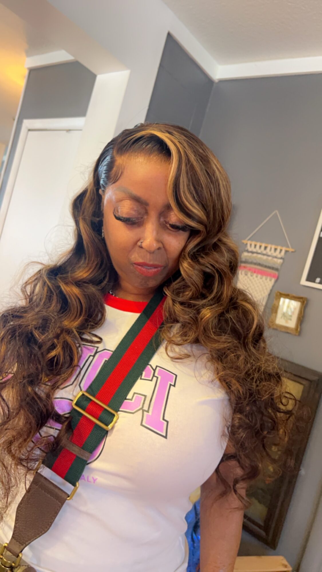 Woman with long, wavy hair wearing a white shirt with red and purple accents, standing indoors with wall decorations.