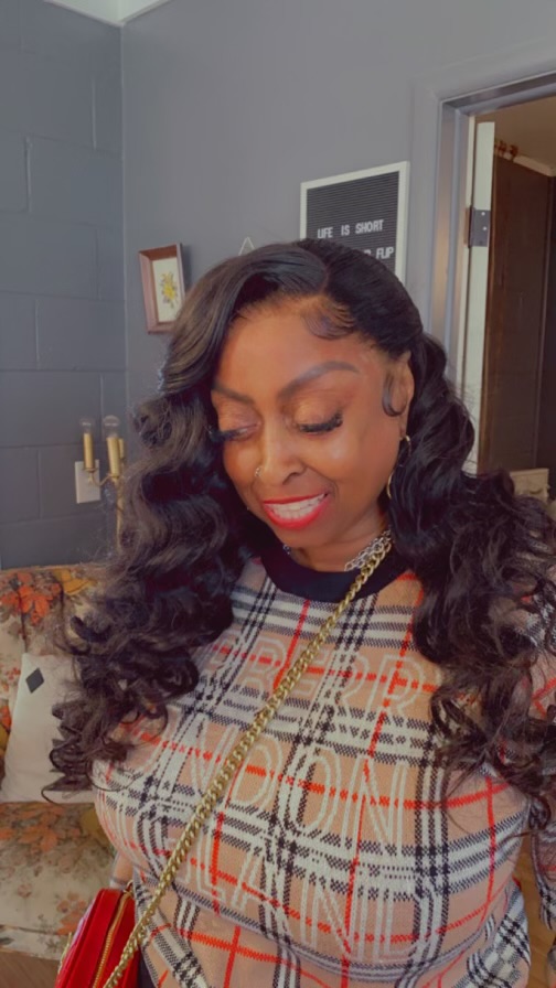 Woman with long, wavy hair smiling, wearing a plaid top and gold necklace, in a room with gray walls and framed pictures.
