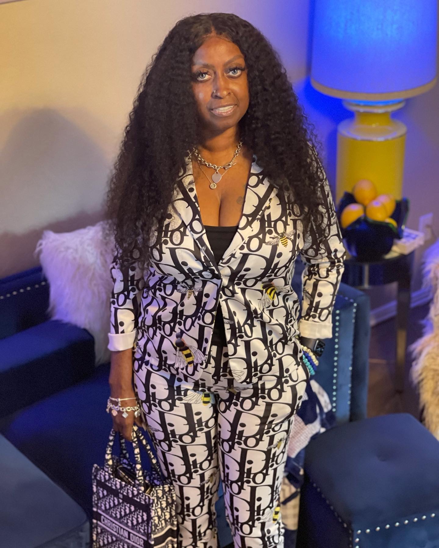 Woman with long curly hair in patterned pajamas holding a handbag, standing in a living room with a lamp and furniture.