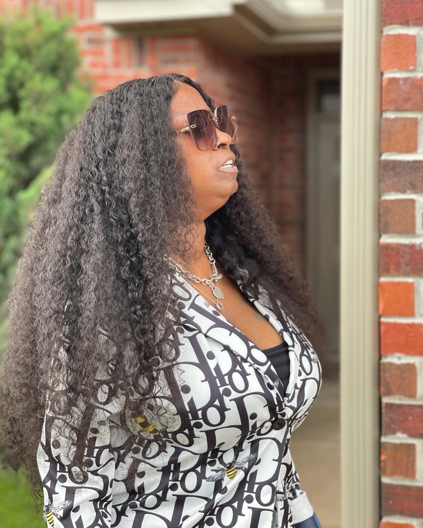 Woman with long curly hair wearing sunglasses and a patterned top standing outside near a brick wall.