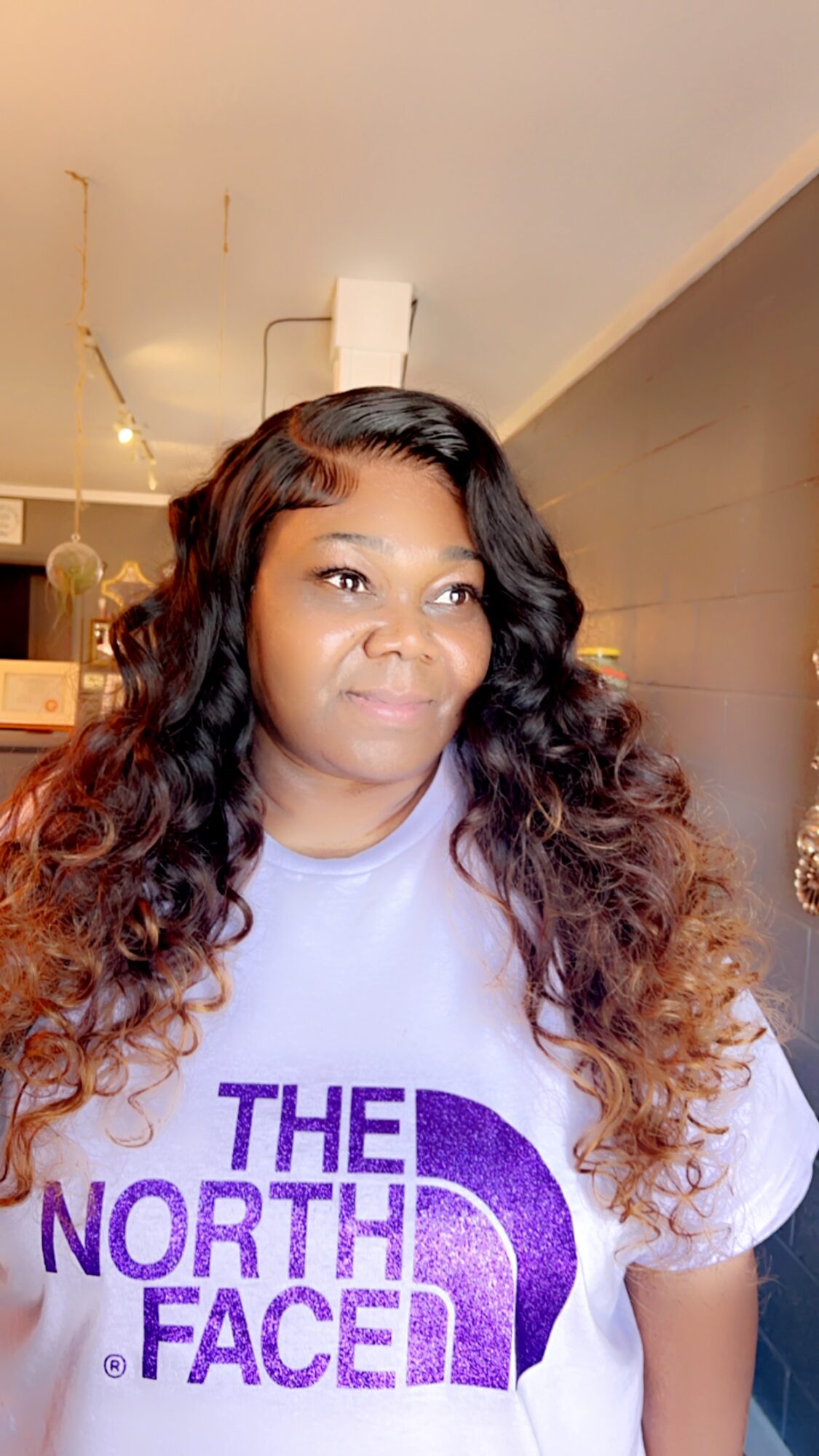 Woman with long, wavy hair wearing a light-colored North Face shirt, standing indoors with a ceiling and wall visible.