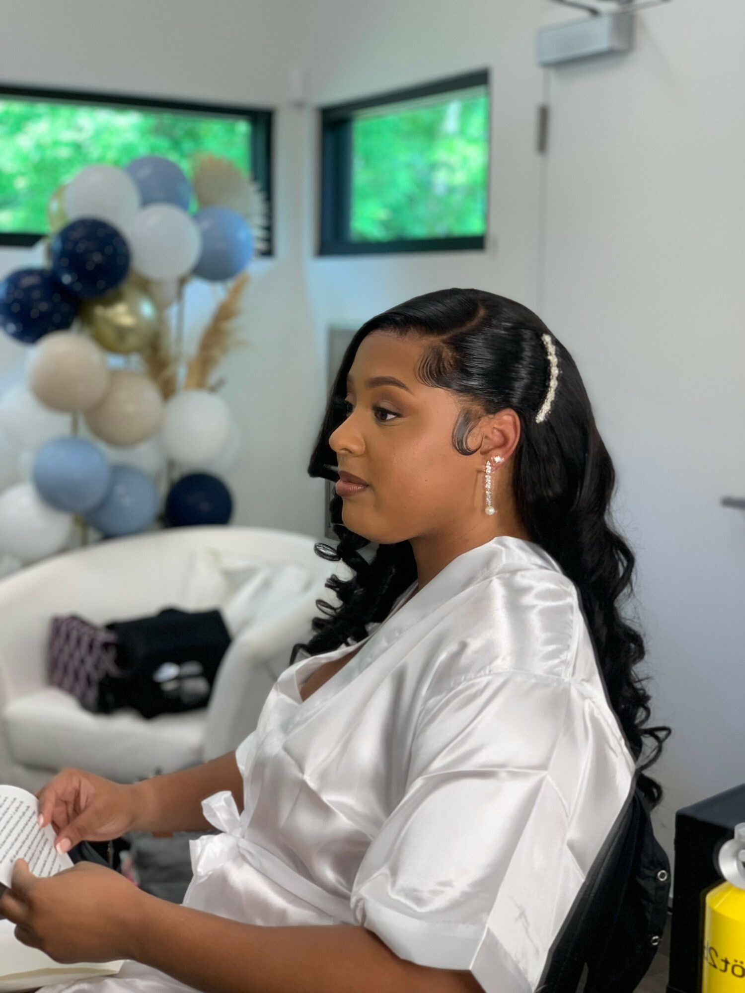 Woman with long wavy hair and pearl hair accessories, wearing a white satin robe, sitting indoors near windows with balloons in the background.
