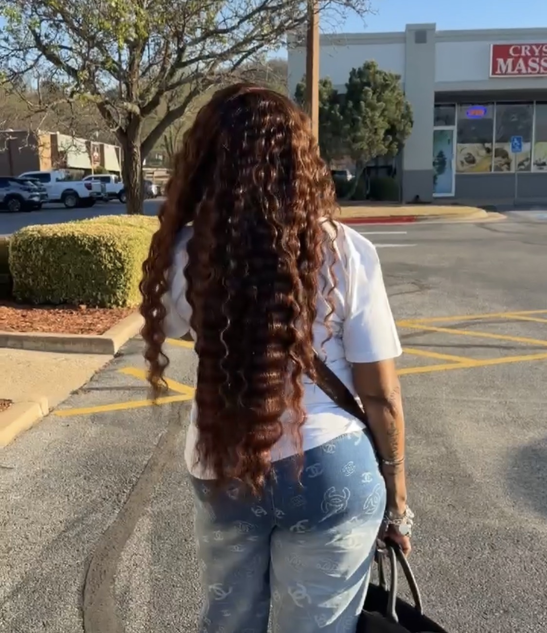Person with long curly hair walking in parking lot, facing away from camera, holding a bag, with trees and building in background.
