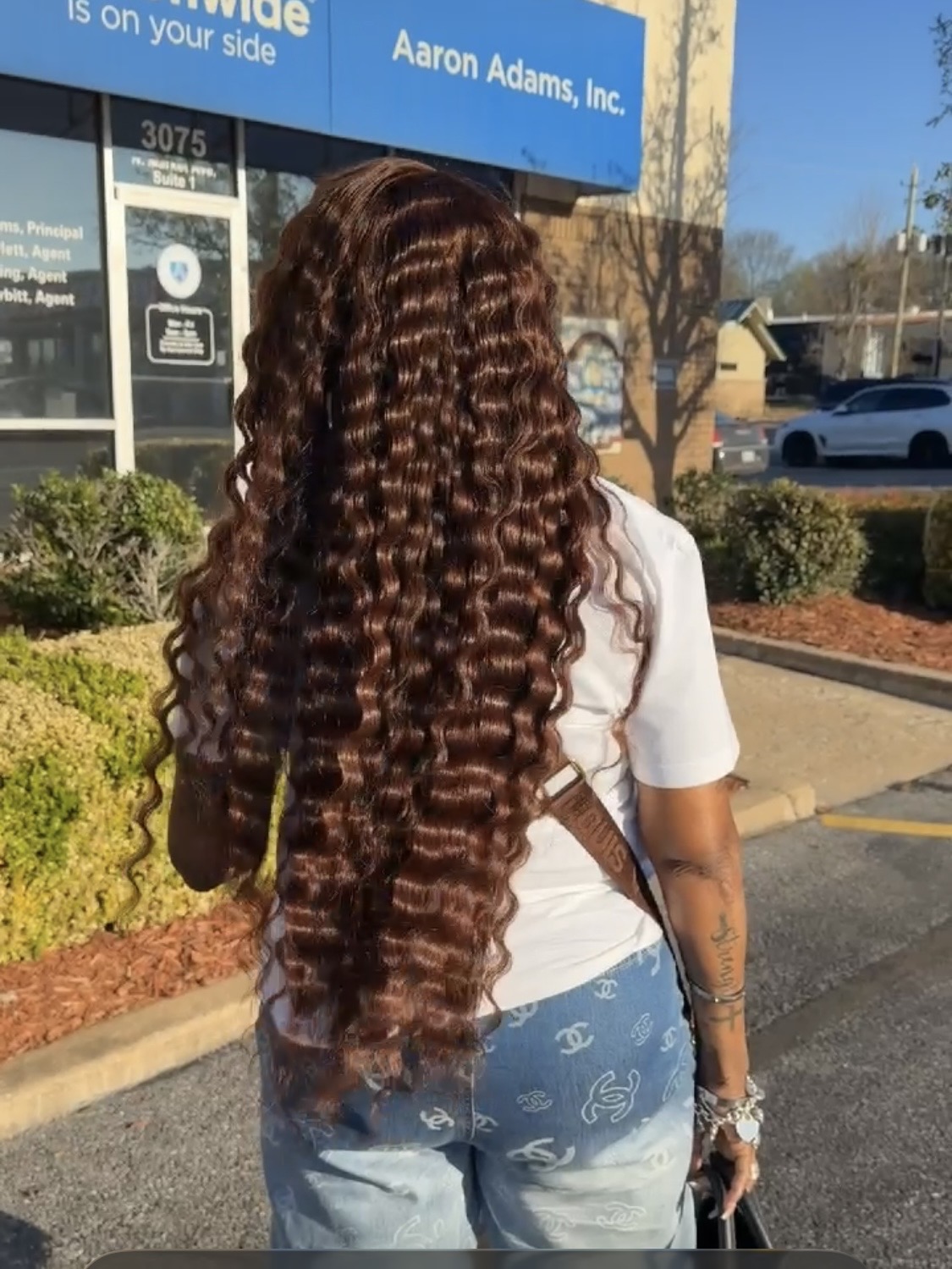Back view of a woman with long, curly hair wearing a white shirt and light blue jeans, standing outdoors.