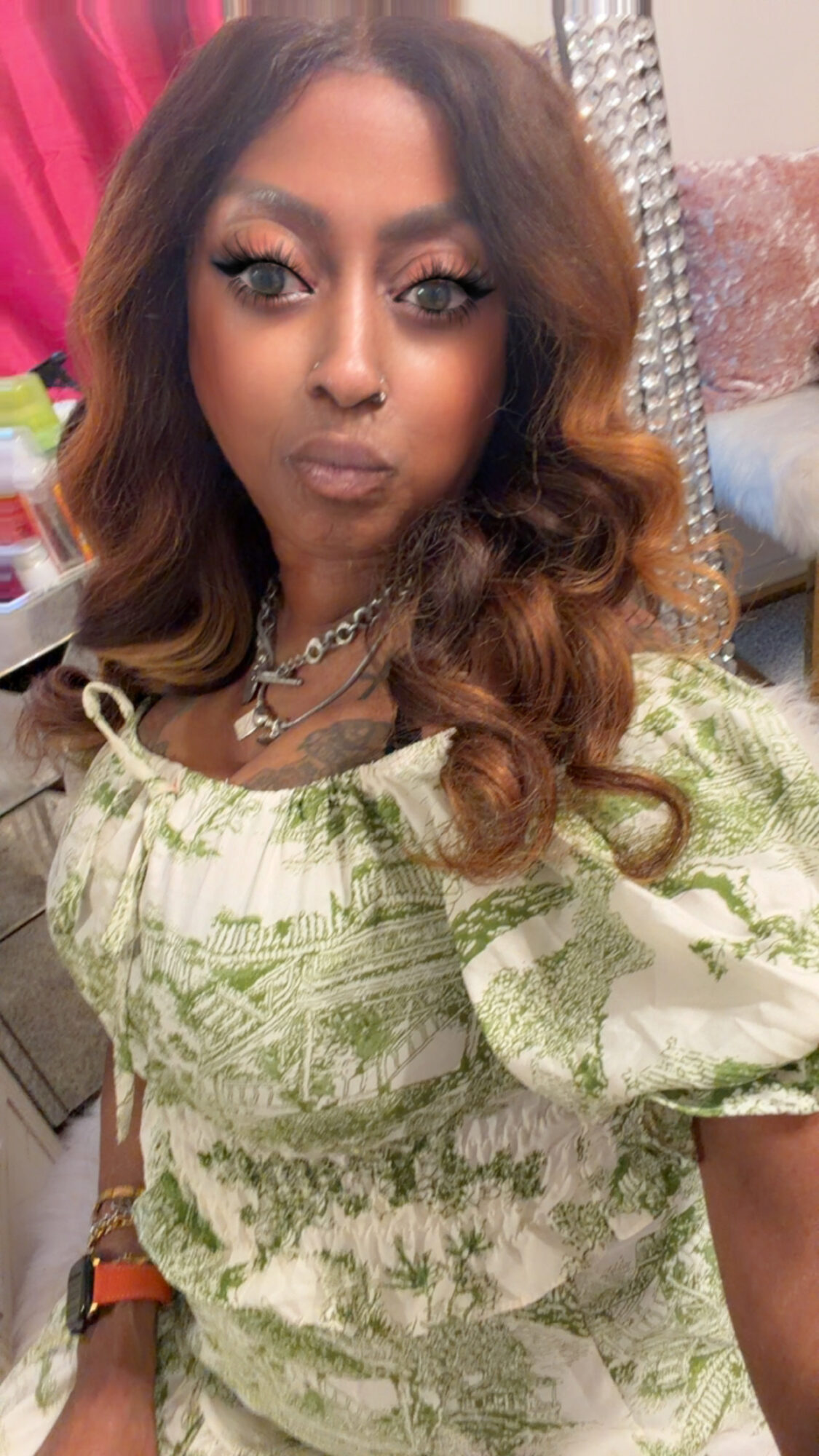 Woman with wavy hair and makeup, wearing a green patterned top, jewelry, and a watch, in an indoor setting.