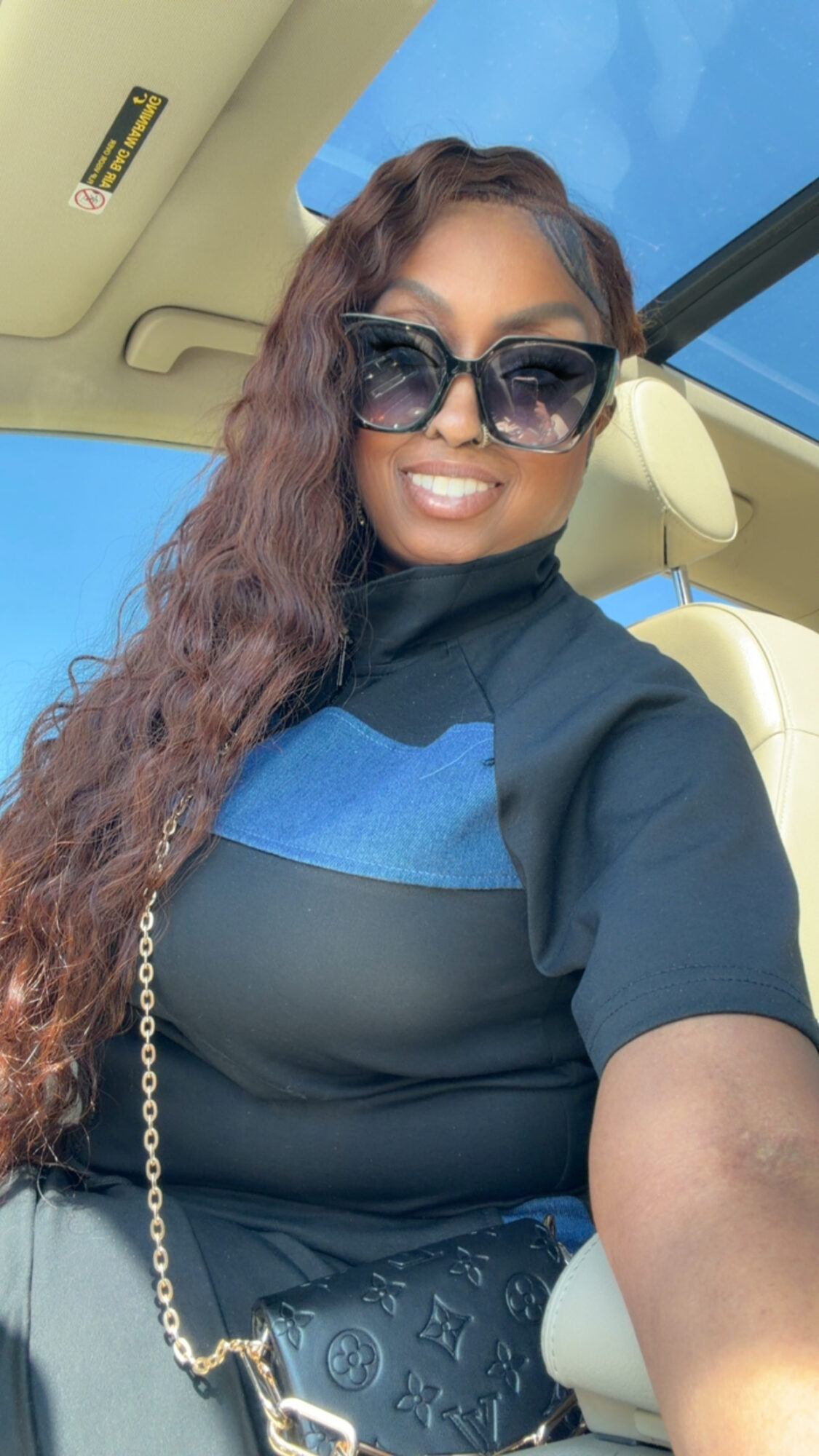 Woman with long wavy hair wearing sunglasses and a dark top, sitting in a car with a blue sky visible through the window.