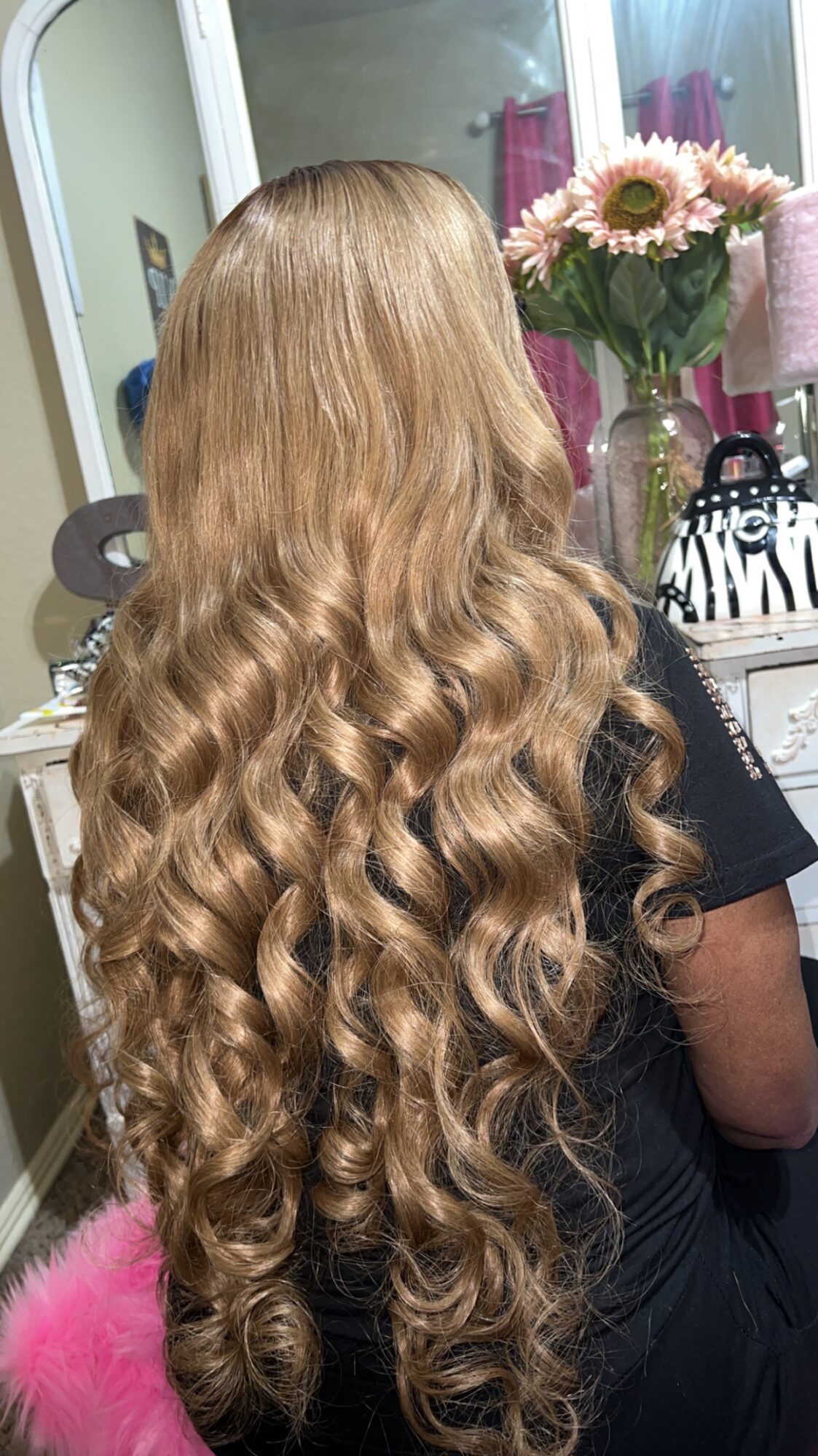 Person with long, wavy, blonde hair in a room with a mirror, flowers, and a dresser, viewed from behind.