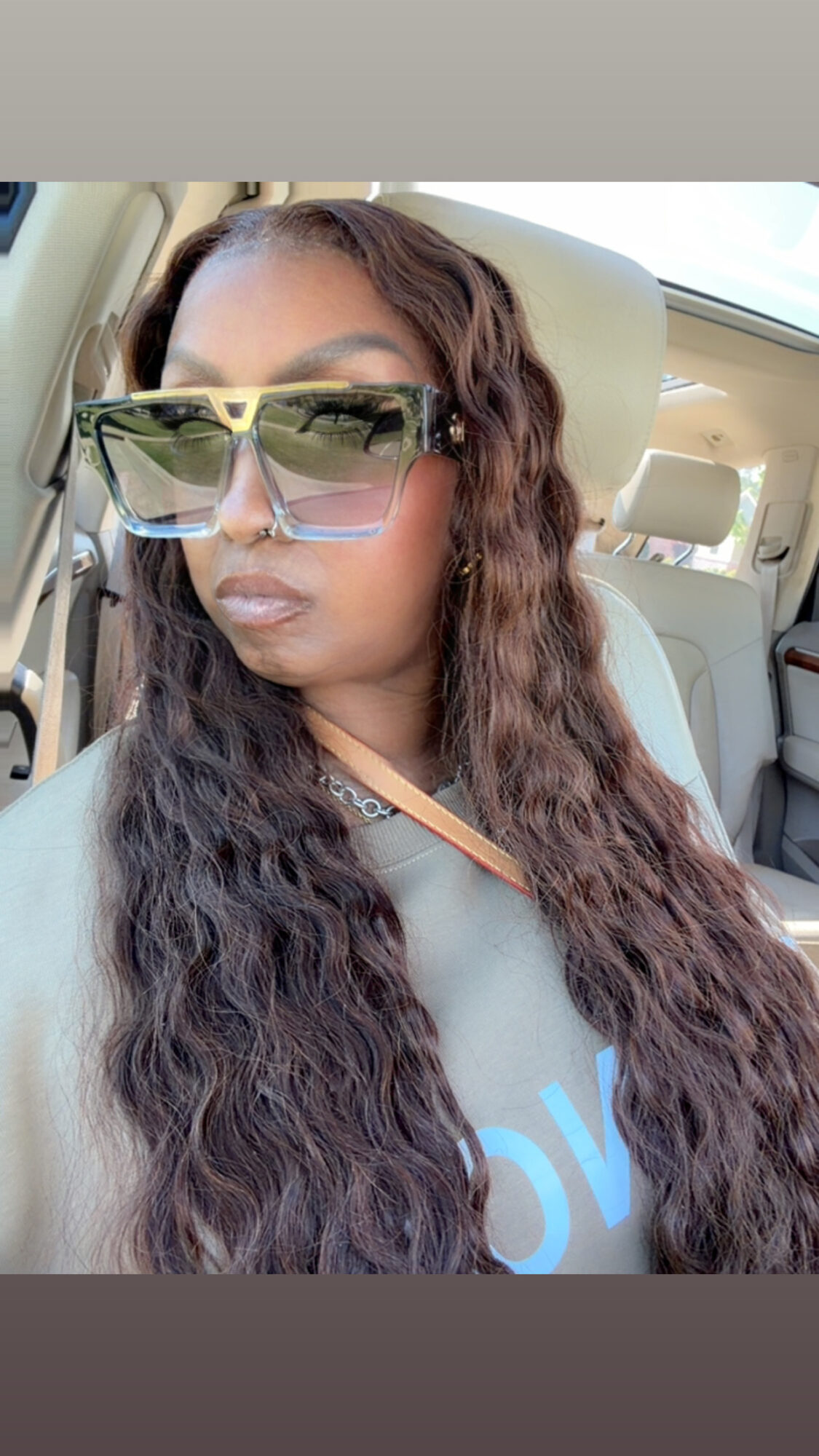 Woman with long wavy hair wearing large sunglasses sitting in a car, sunlight visible through window