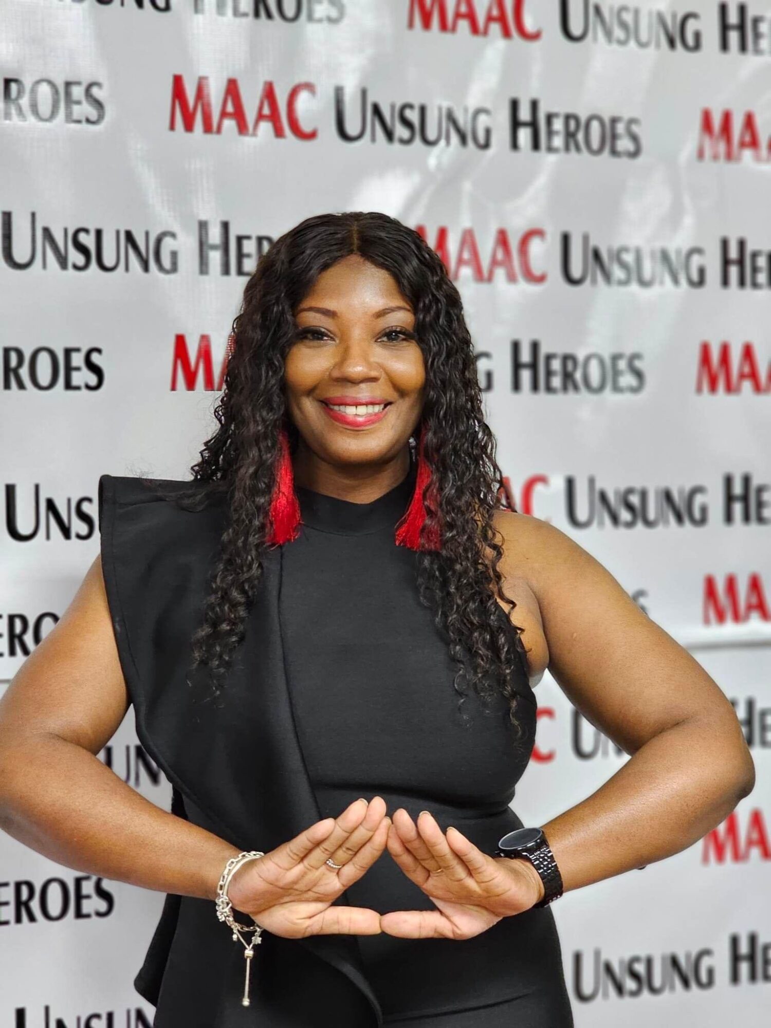Woman smiling with curly hair, wearing black sleeveless top, red earrings, and a watch, standing in front of a backdrop.